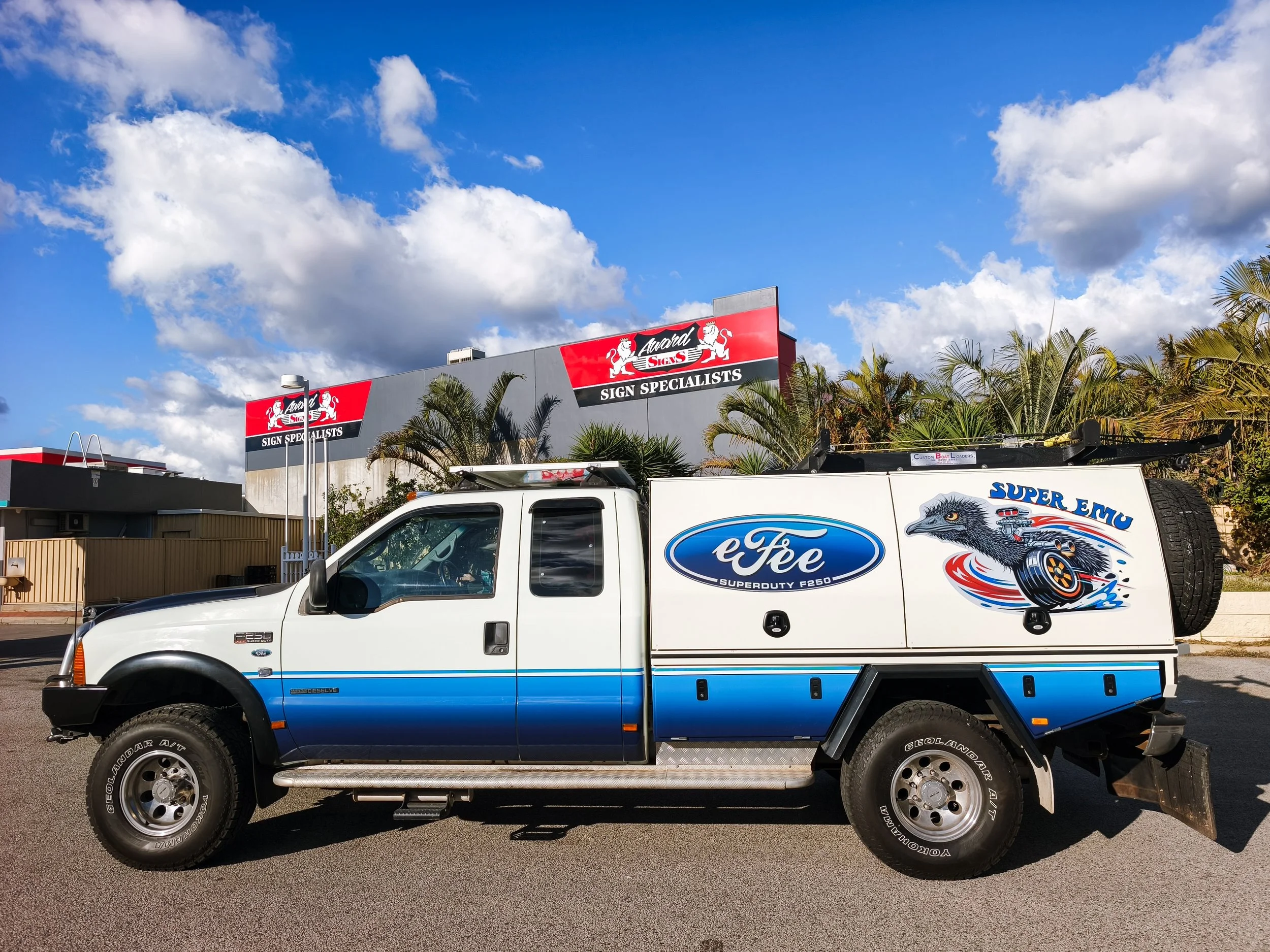 A white and blue pickup truck parked near a sign for 'Award Sign Specialists' under a partly cloudy sky. The truck has a graphic of a dragon and racing elements on its side.