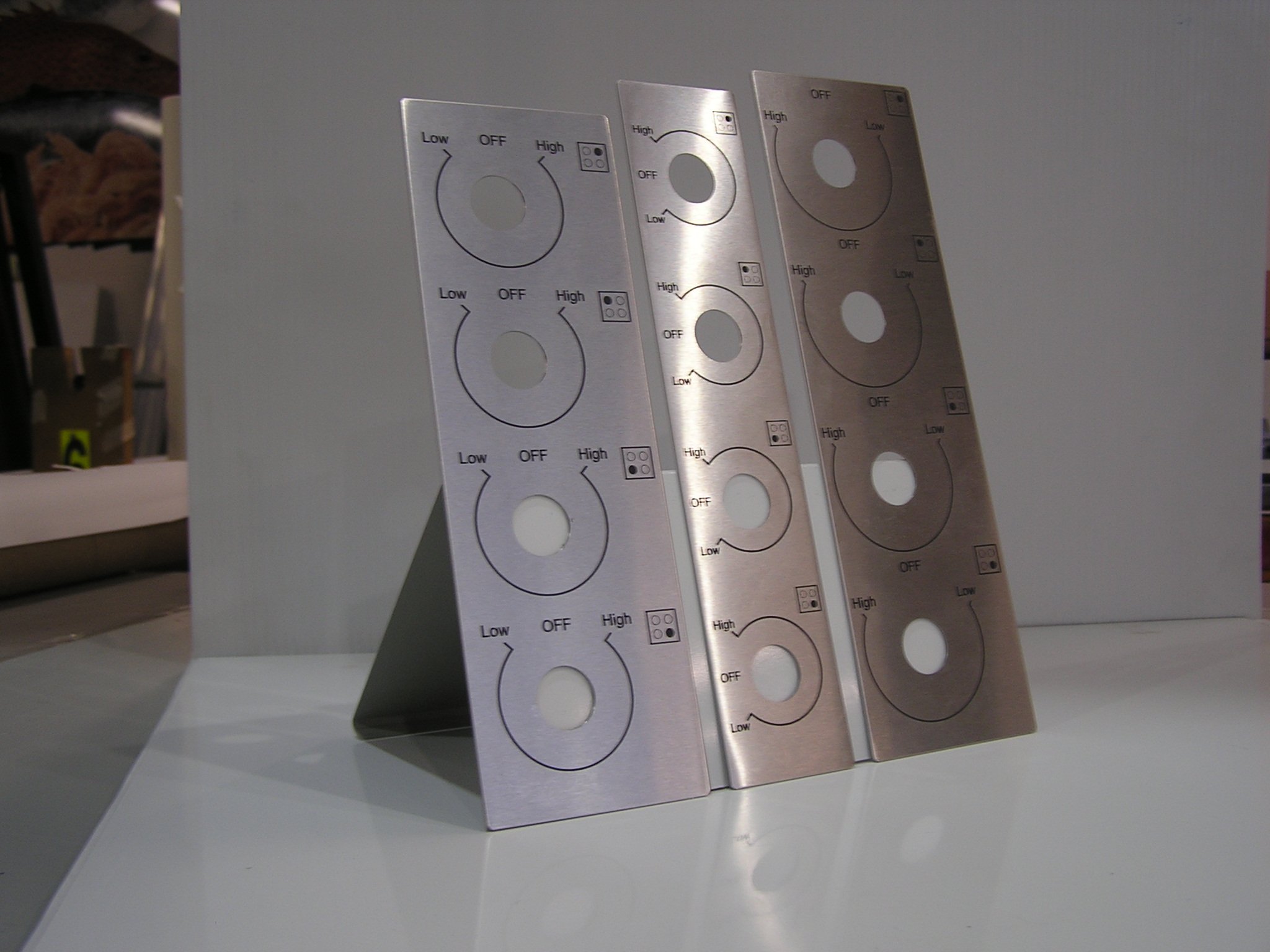 Close-up of three metallic control panels with circular dial labels for light dimming, mounted on a white wall.