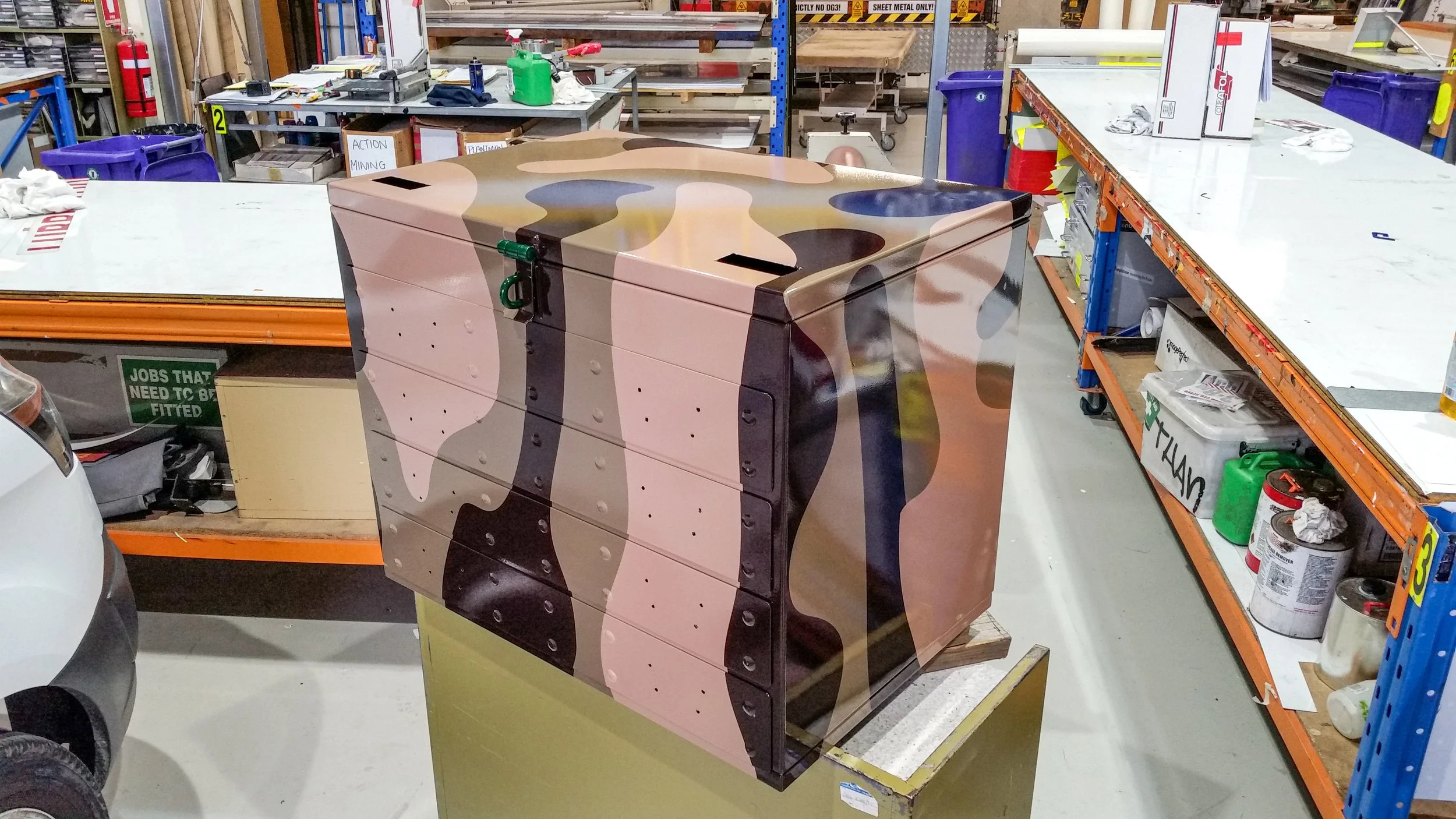A storage cabinet with a camouflage pattern in a workshop or warehouse setting, against a background of shelves, worktables, and various tools and supplies.