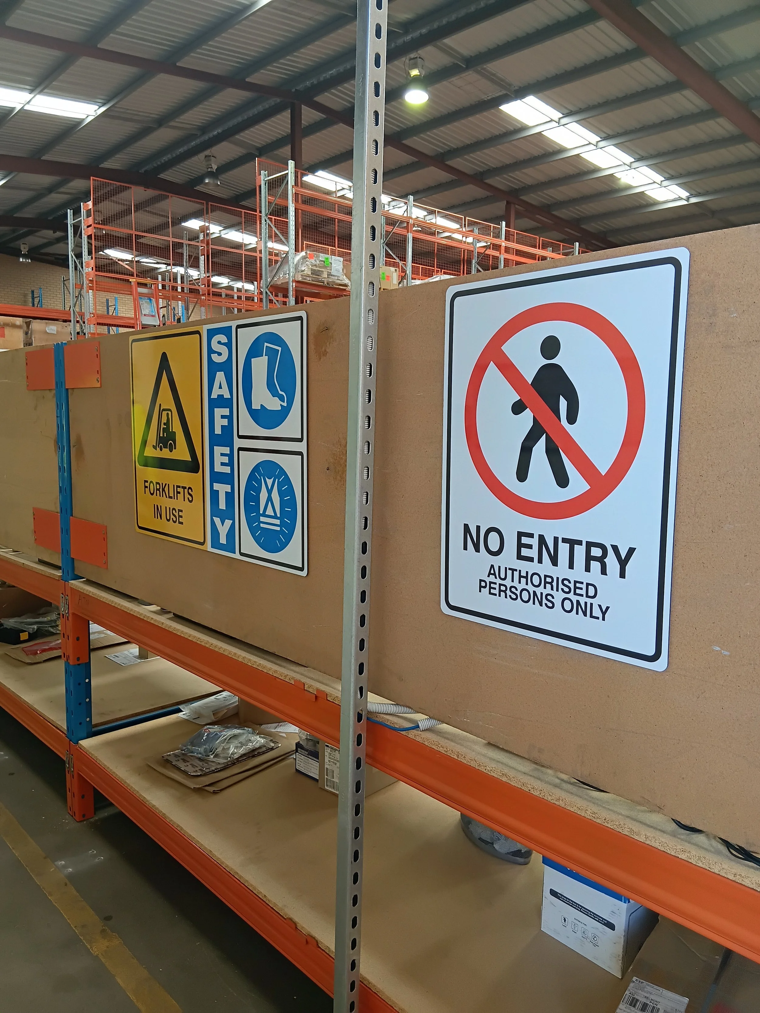 Warehouse aisle with metal shelving and safety signs, including a warning about forklift use, safety equipment, and no entry for unauthorized persons. Mandatory Safety Signage