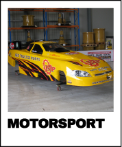 Yellow custom drag racing car with black and red fire patterns and 'RACECRAFT POWER' written on the side, positioned inside a garage with shelves and storage containers in the background.