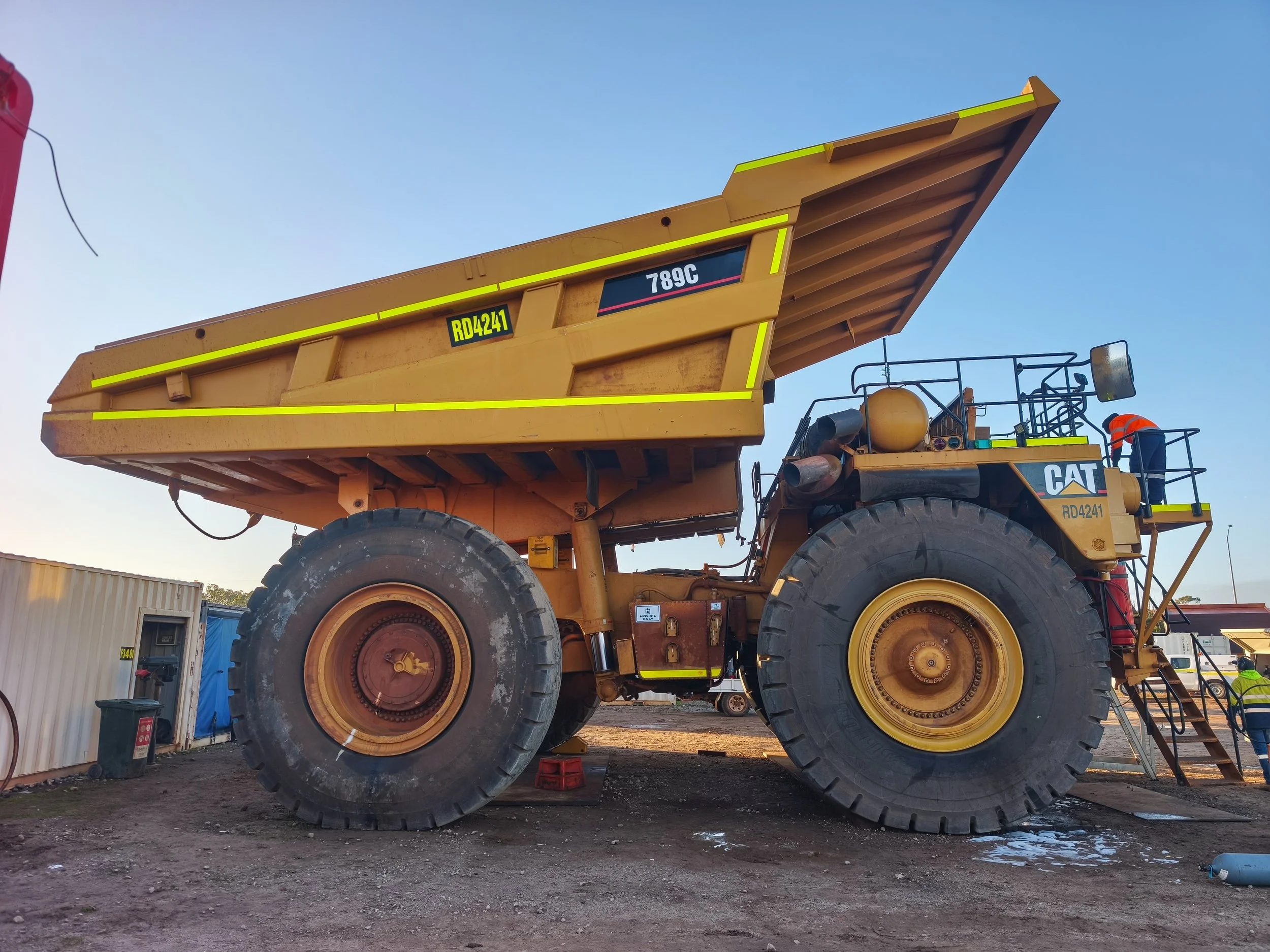 A large yellow Caterpillar industrial dump truck parked outdoors with clear blue skies in the background. Two workers in safety vests and helmets are near the truck, and various equipment and buildings are visible around the site.