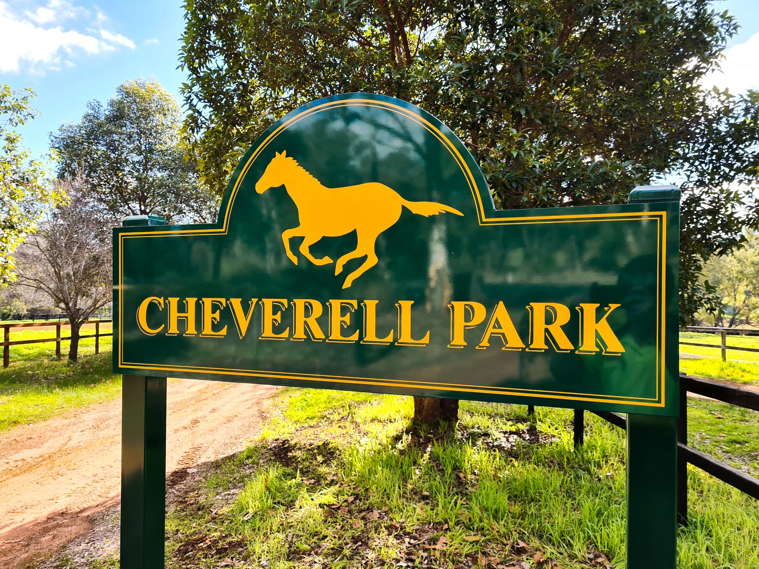 Sign for Cheverell Park with a yellow horse logo on a green background, outdoors with trees and a dirt pathway in the background. Business entrance road street signage and branding
