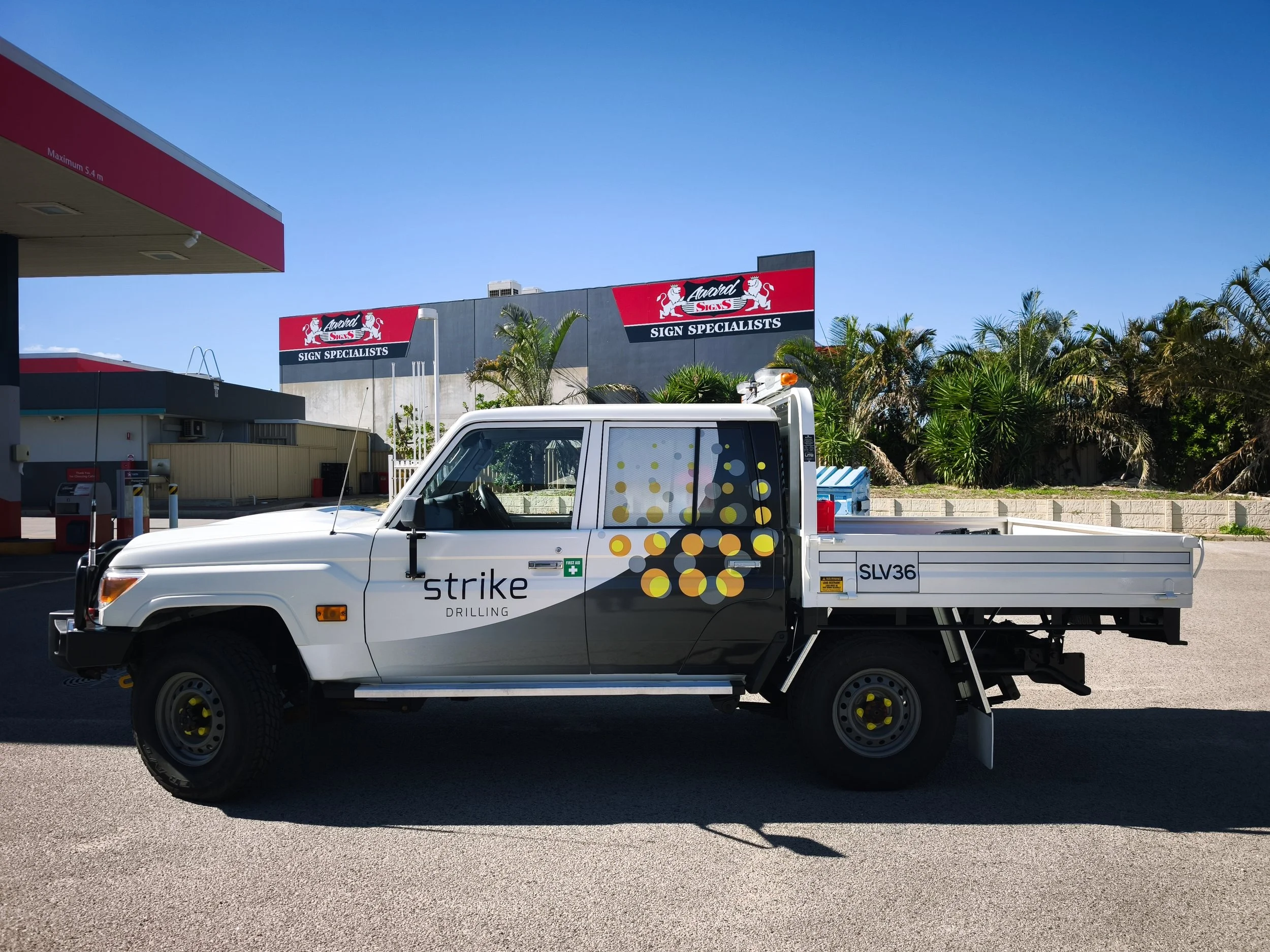 A white flatbed truck with yellow and black wheels parked in an area with palm trees, near a sign and a building. The sign reads 'Award Signs Sign Specialists.'
