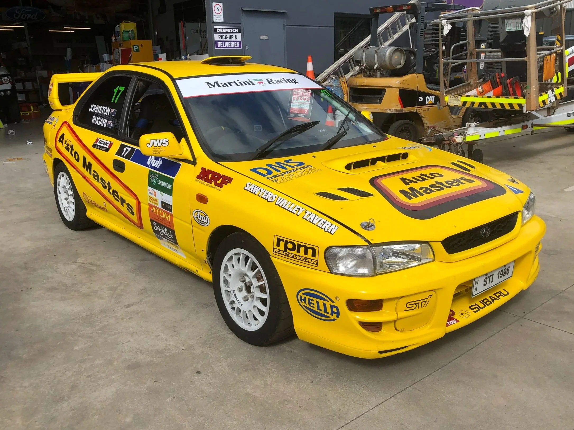 A bright yellow GC8 Subaru WRX STI Rally Racecar with a racing number and features logos such as Auto Masters, RPM Racewear, and Subaru. Motorsport sponsors