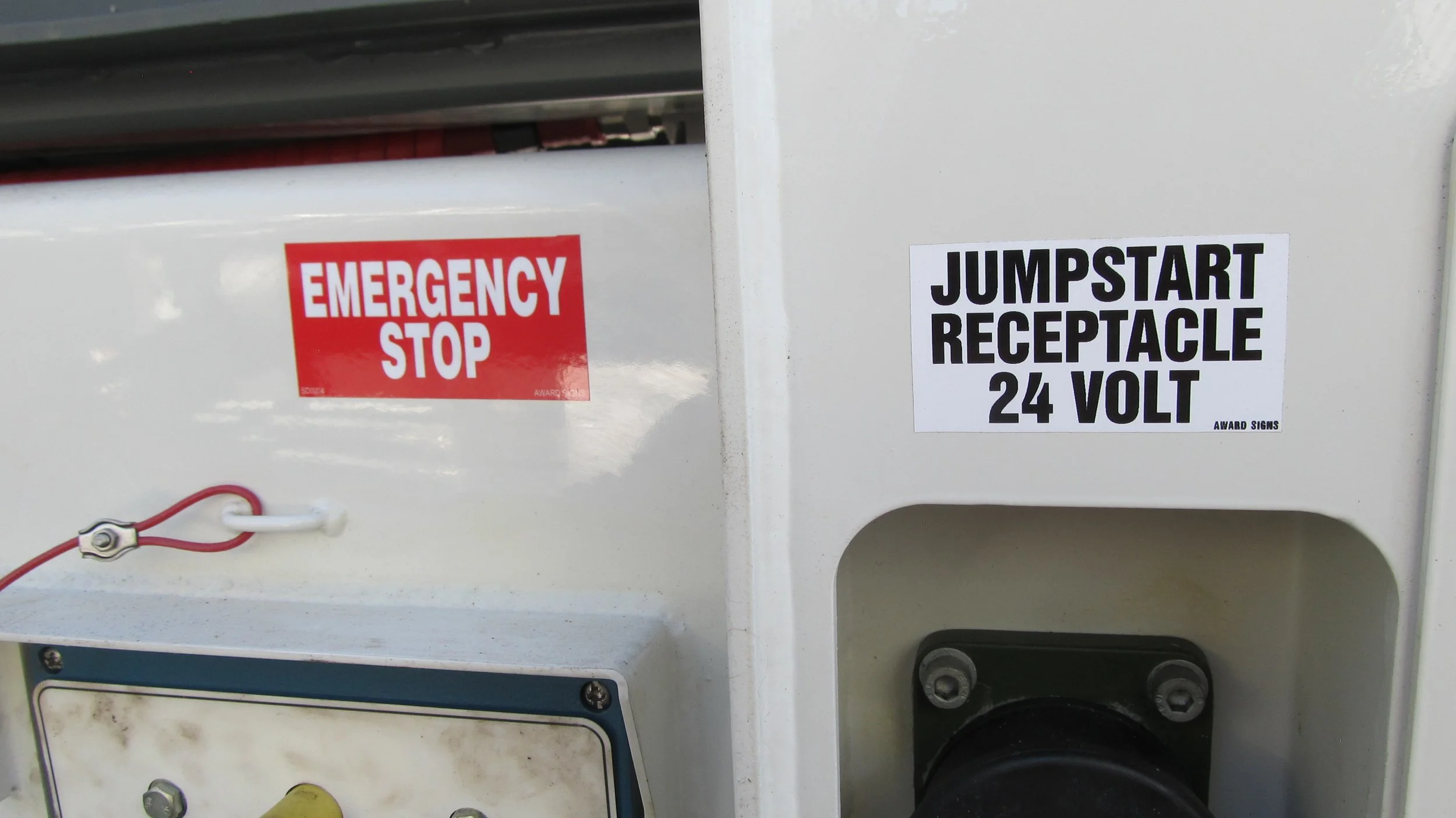 Close-up of two signs on electrical equipment. The first sign is red and white, reading 'EMERGENCY STOP'. The second sign is white with black text, reading 'JUMPSTART RECEPTACLE 24 VOLT'.
