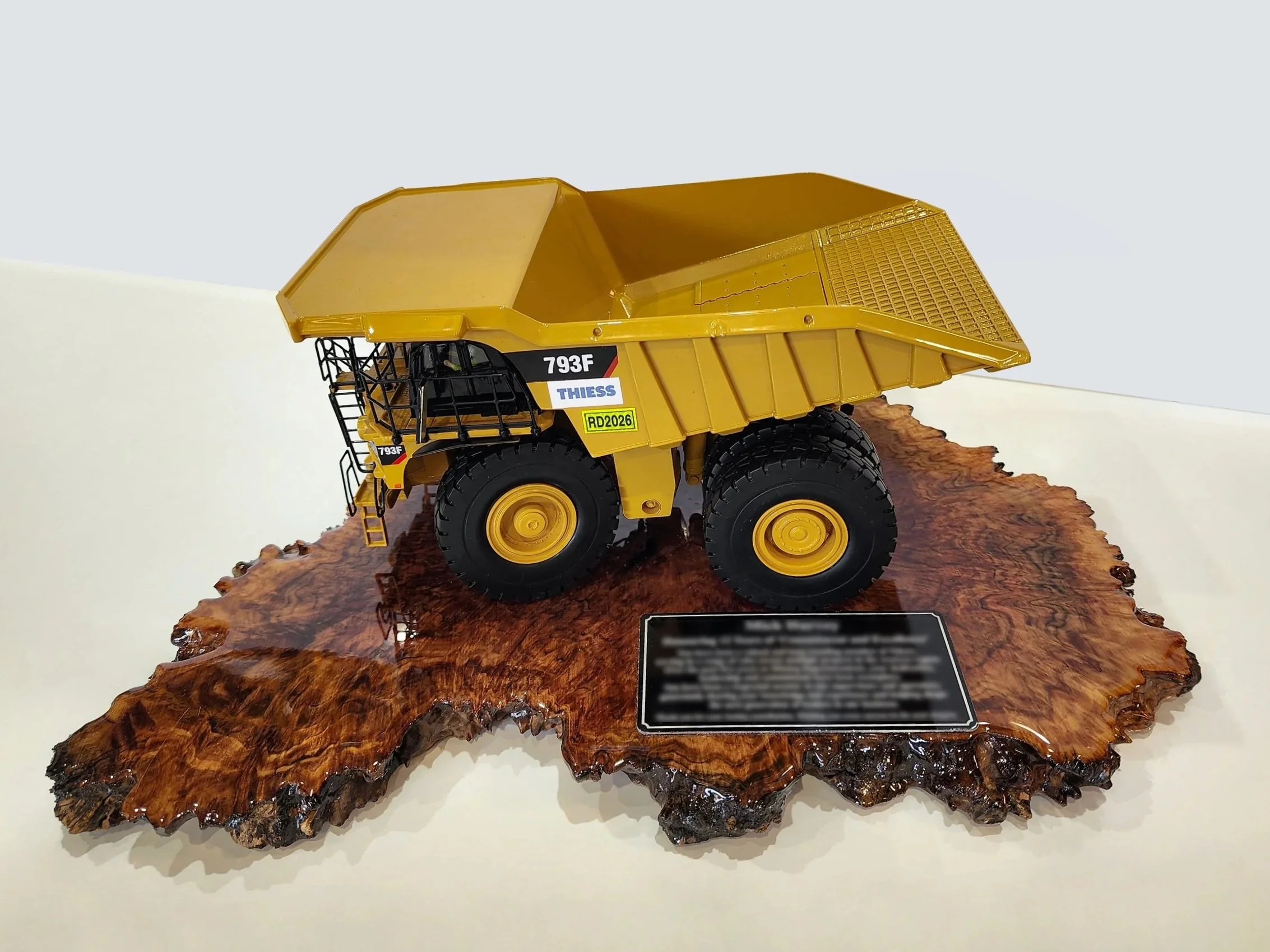 A yellow toy dump truck with the labels "793F," "THIESS," and "RD2026" on its side, placed on a polished, irregularly shaped wooden base. Custom mining gifts