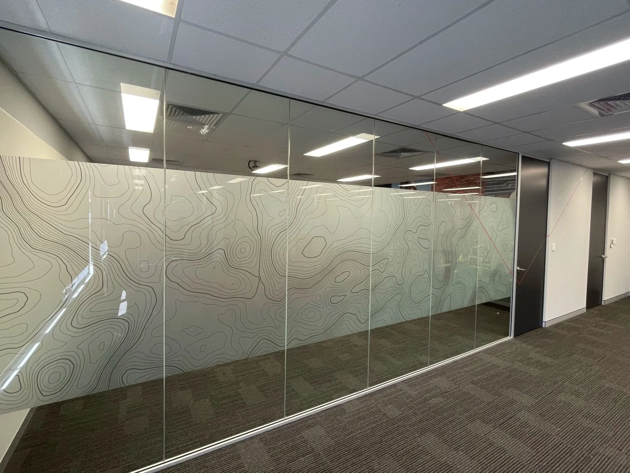 Interior office space with glass wall frosting featuring a topographical map design, ceiling tiles, fluorescent lighting, and a door on the right.