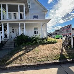 A white two-story house with a porch, surrounded by a lawn and bushes, with a sidewalk in front and a sign on the lawn.