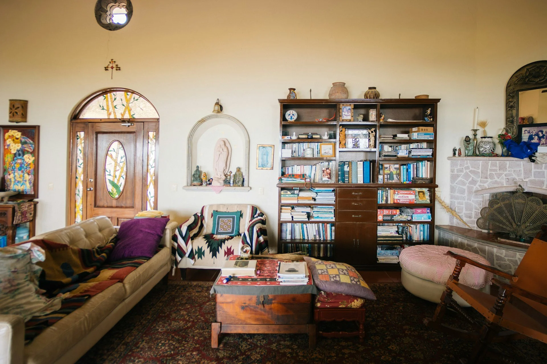 Living room with a beige sofa, a colorful armchair, a wooden coffee table, a large wooden bookshelf filled with books, various decorative items, a stone fireplace, and a wooden door with stained glass windows.