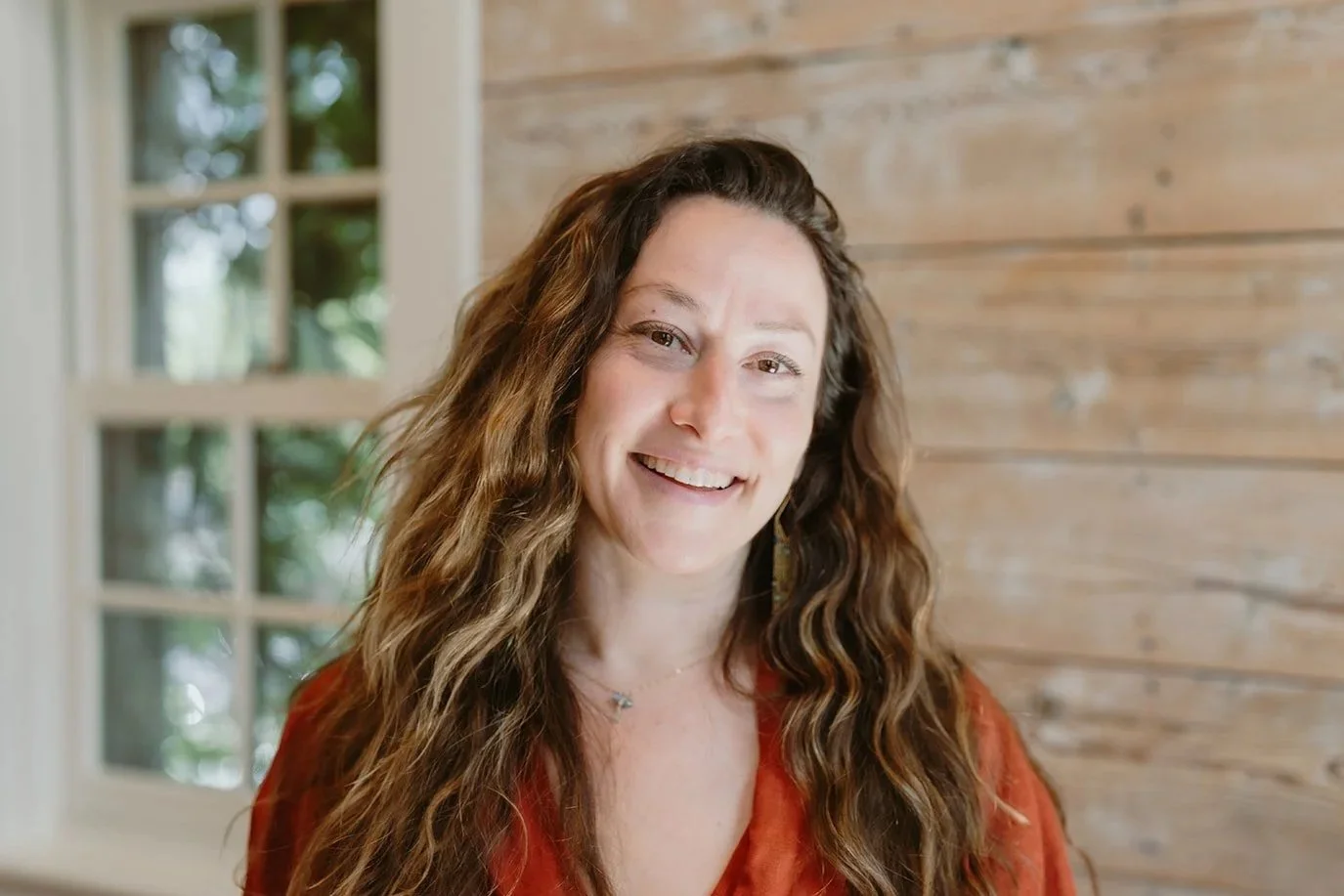 A woman with long curly brown hair smiling in front of a window and wooden wall.