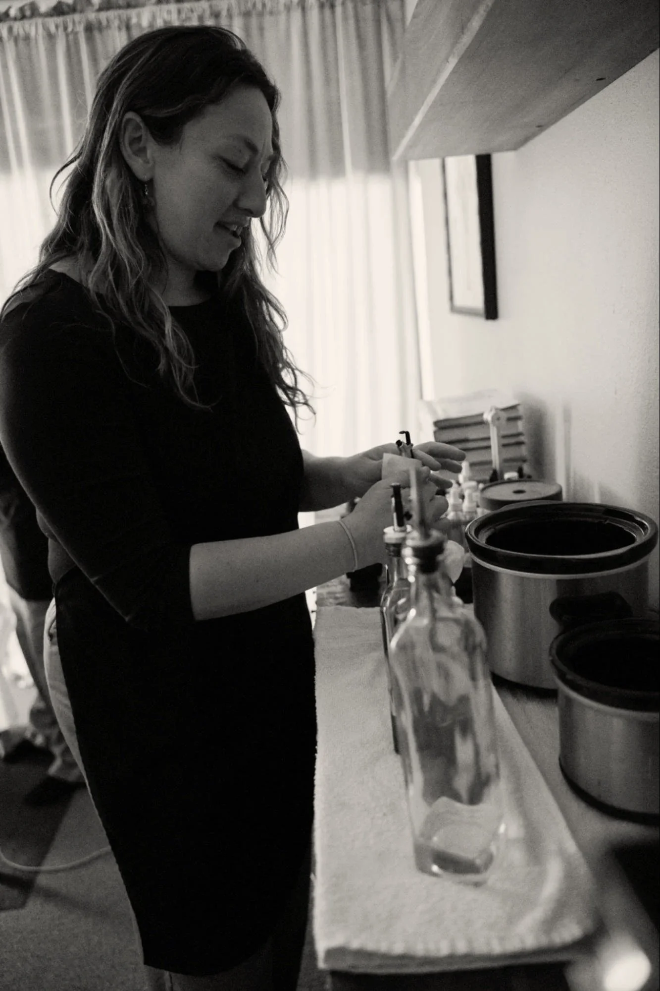 A woman with wavy hair wearing a dark shirt is working in a kitchen, handling a syringe and other tools on the counter.