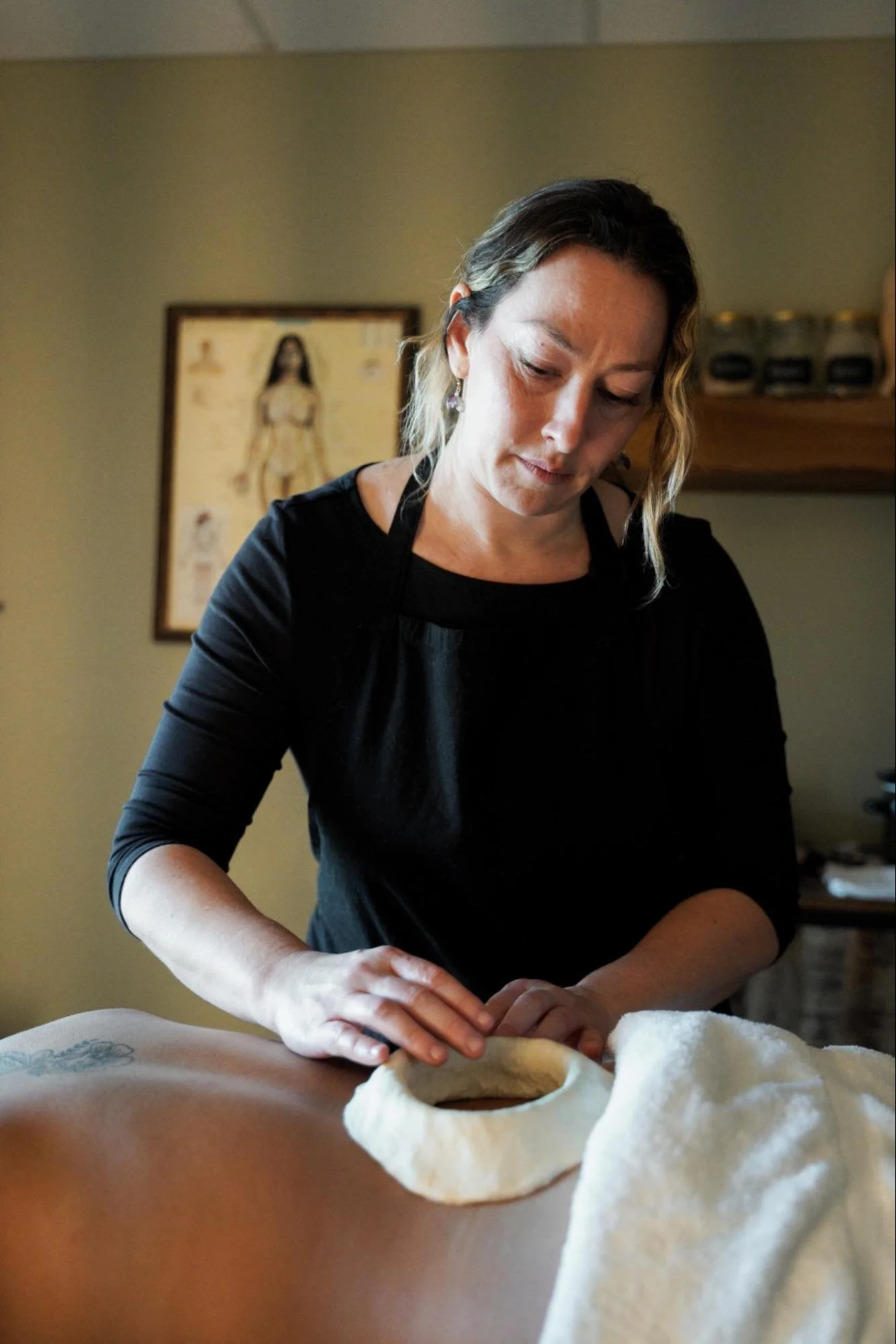 Woman with wavy hair wearing earrings rolling dough on a table in a kitchen with a tattoo in the background.