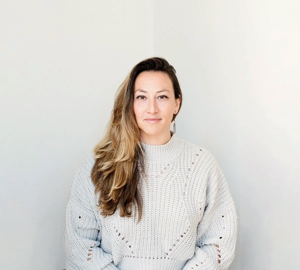 A woman with long, wavy brown hair, wearing a cream knitted sweater, standing in front of a plain white wall.