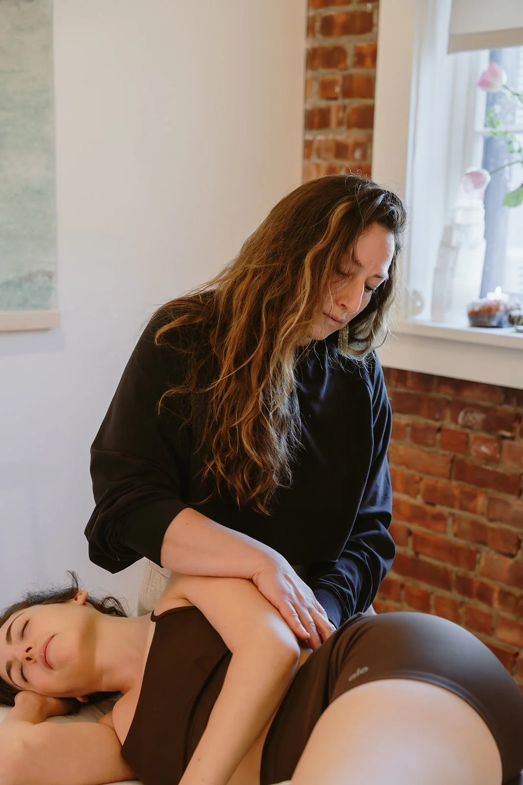 A woman giving a massage to another woman who is lying down on a bed or table in a cozy room with exposed brick wall and a window in the background.