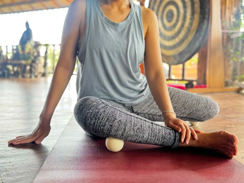 A person practicing yoga or stretching indoors, sitting cross-legged on a yoga mat with a massage or therapy ball under their leg.