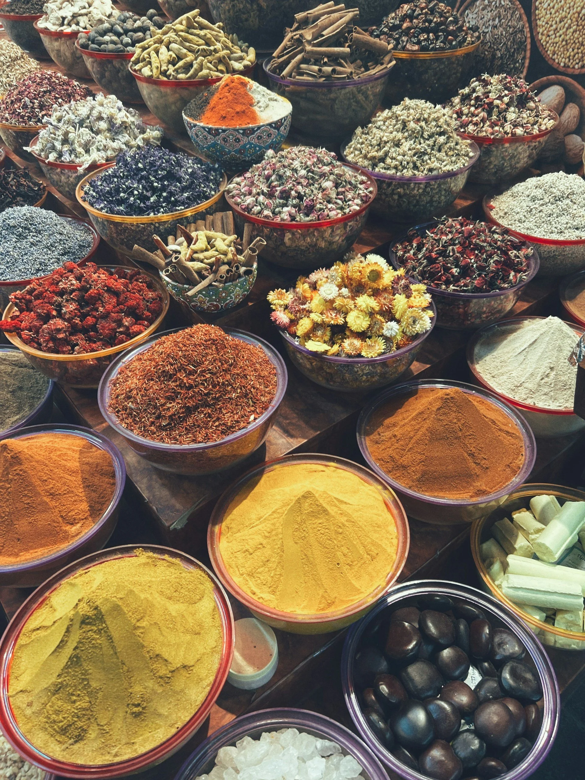 Colorful spices and herbs in bowls at a market stall, including yellow turmeric, red chili powder, dried flowers, cinnamon sticks, dried herbs, black stones, and white blocks.