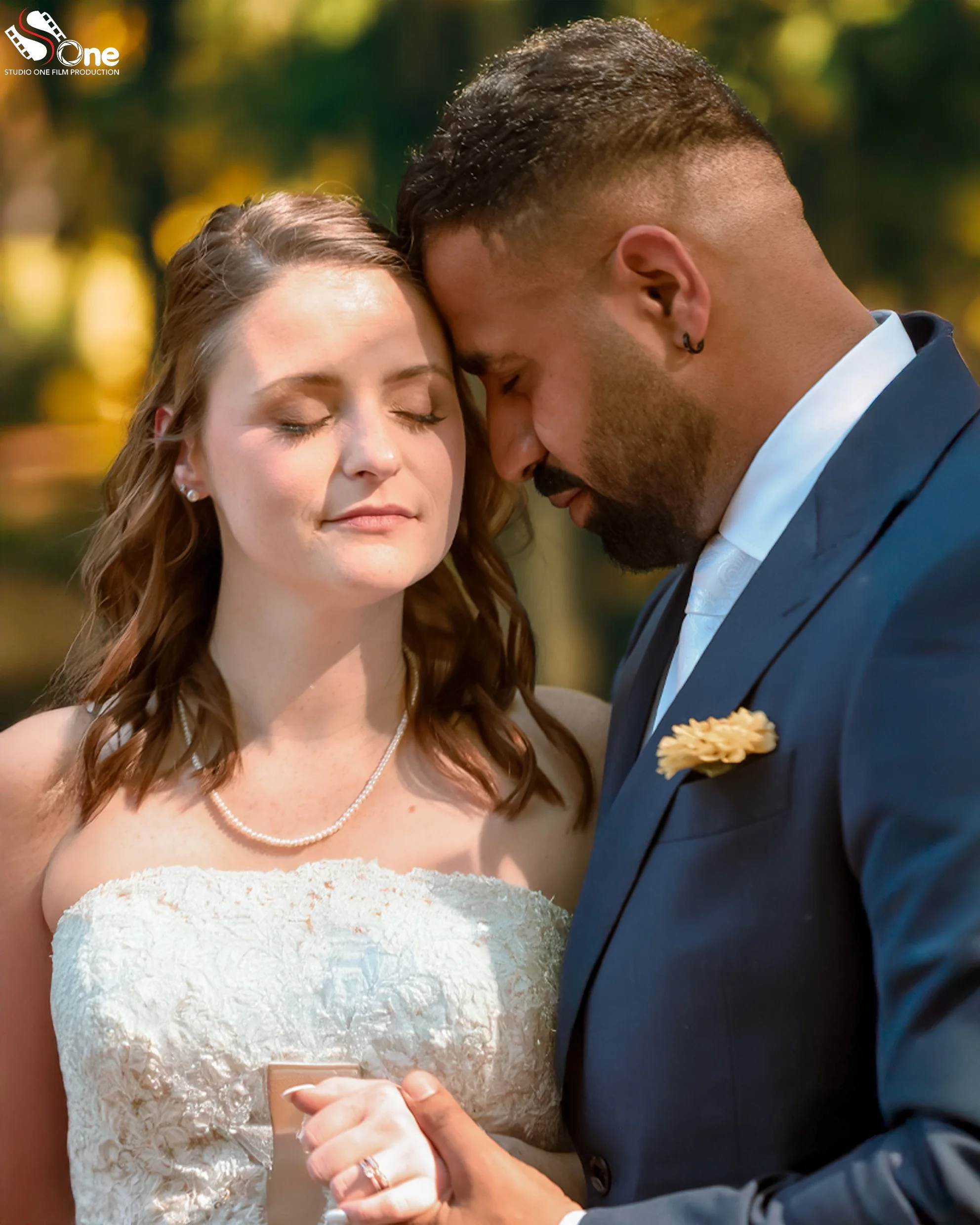 A bride and groom with foreheads touching, eyes closed, during their outdoor wedding ceremony, holding hands, with a blurred autumn background.