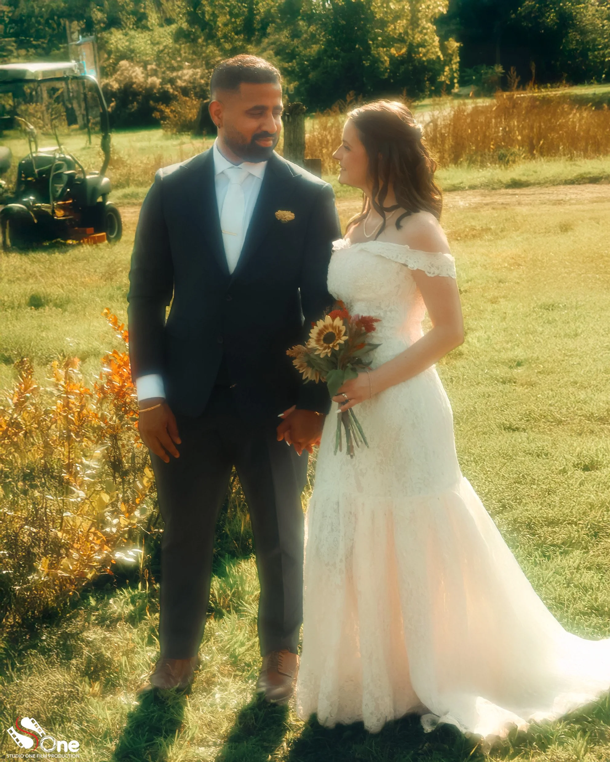 A newlywed couple holding hands outdoors with a grassy field, trees, and a golf cart in the background. The groom wears a dark suit and white shirt, and the bride wears a white lace wedding dress holding a sunflower bouquet.