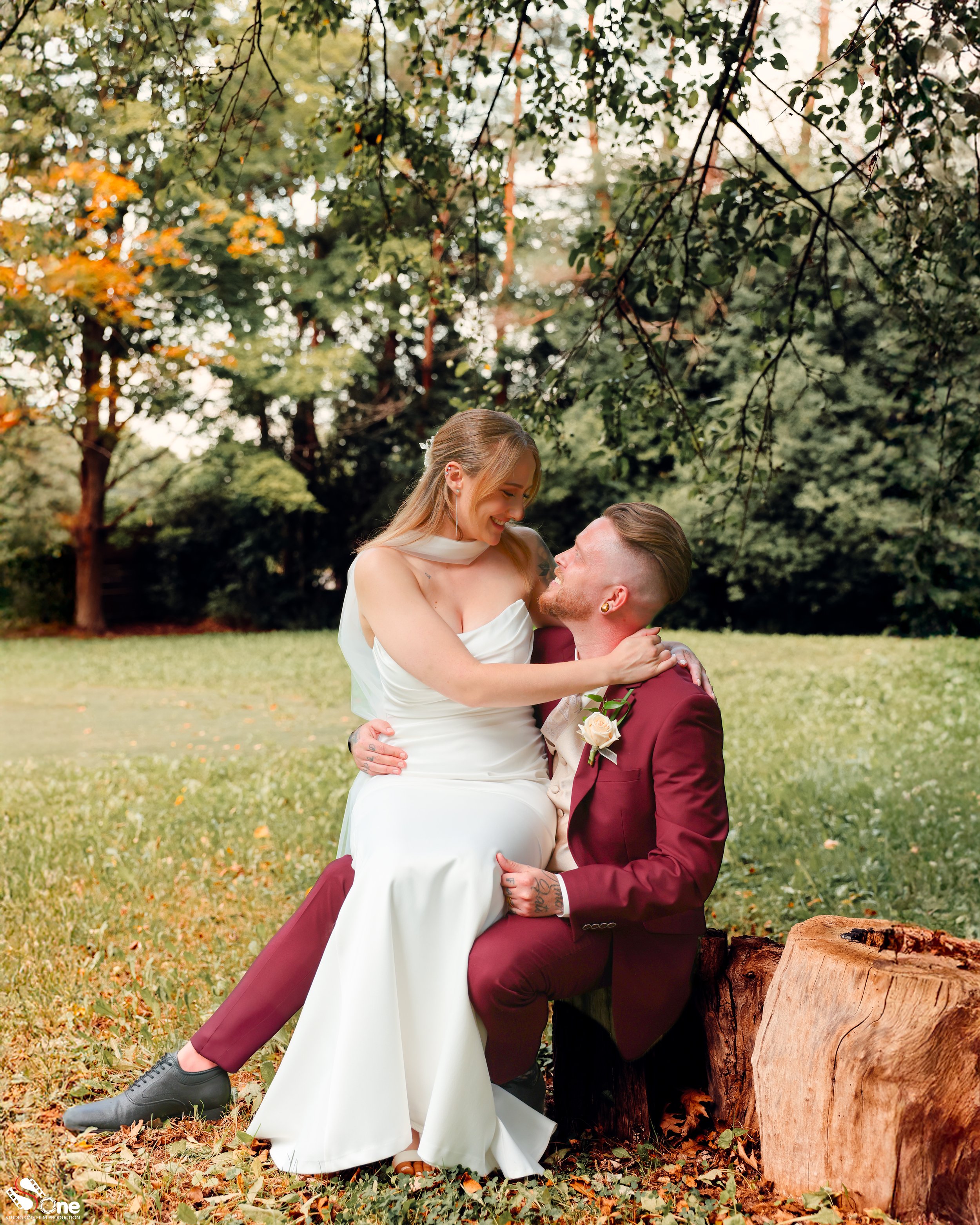 A couple in wedding attire sitting outdoors in a park with green trees in the background. The woman is in a white wedding dress, and the man is in a maroon suit. They are smiling and gazing at each other, with the woman sitting on the man's lap, and he is sitting on a tree stump.