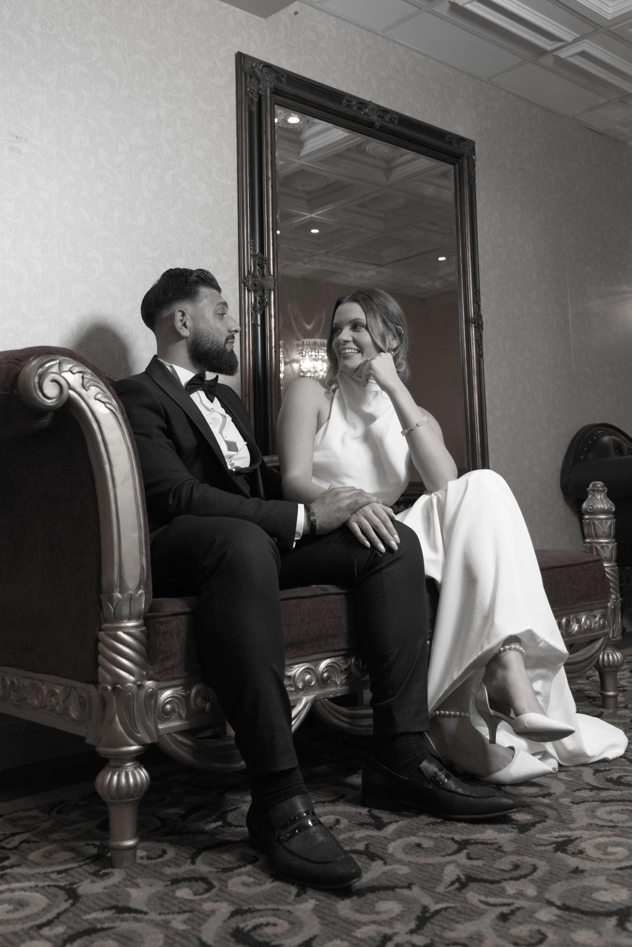 A black-and-white photo of a man in a tuxedo and a woman in a white dress sitting on an ornate sofa, looking at each other and smiling in a luxurious room with a large mirror behind them.