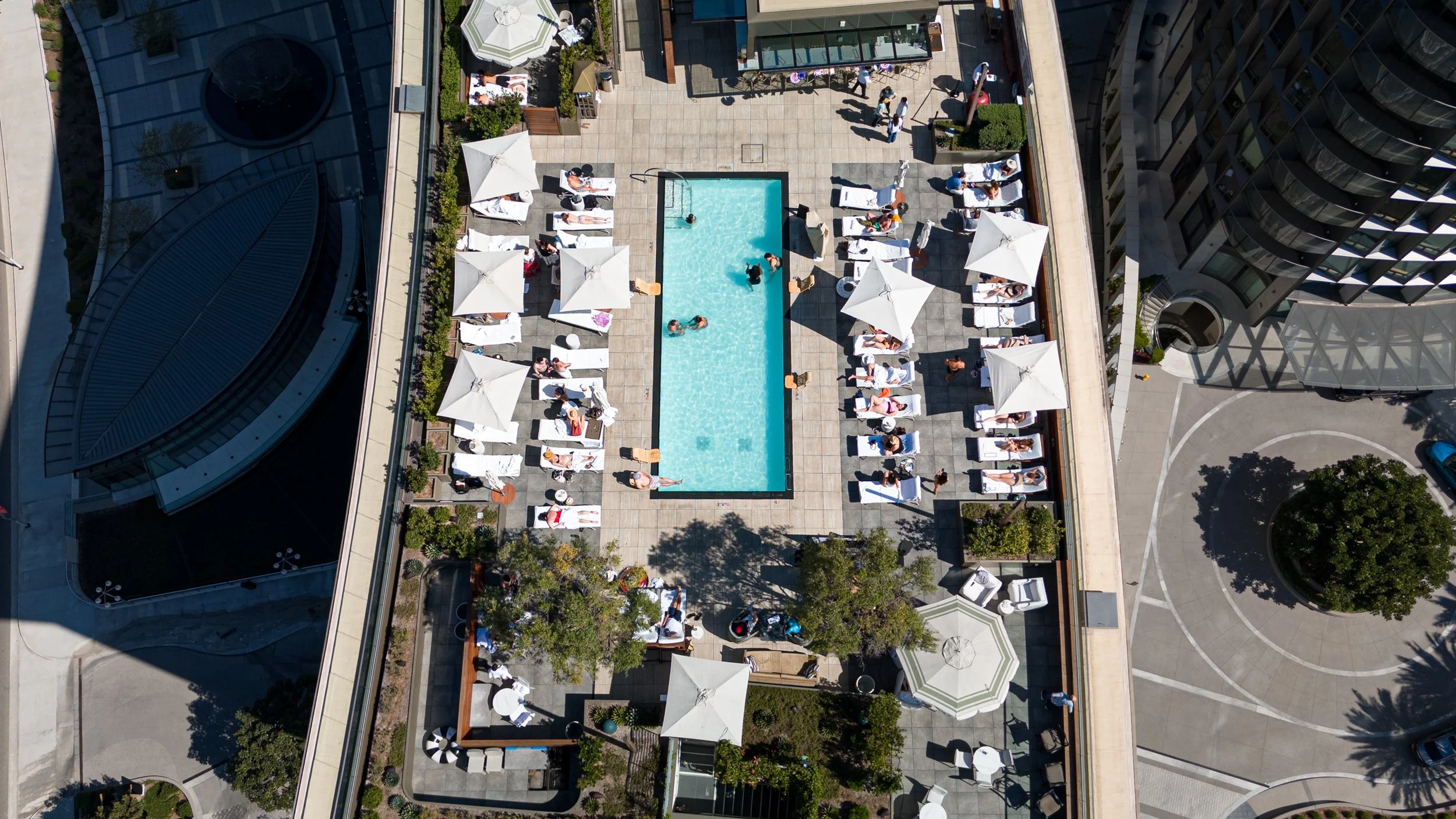 Aerial view of a pool area on a rooftop with people sunbathing under umbrellas and swimming in the pool, surrounded by lounge chairs, umbrellas, and high-rise buildings.