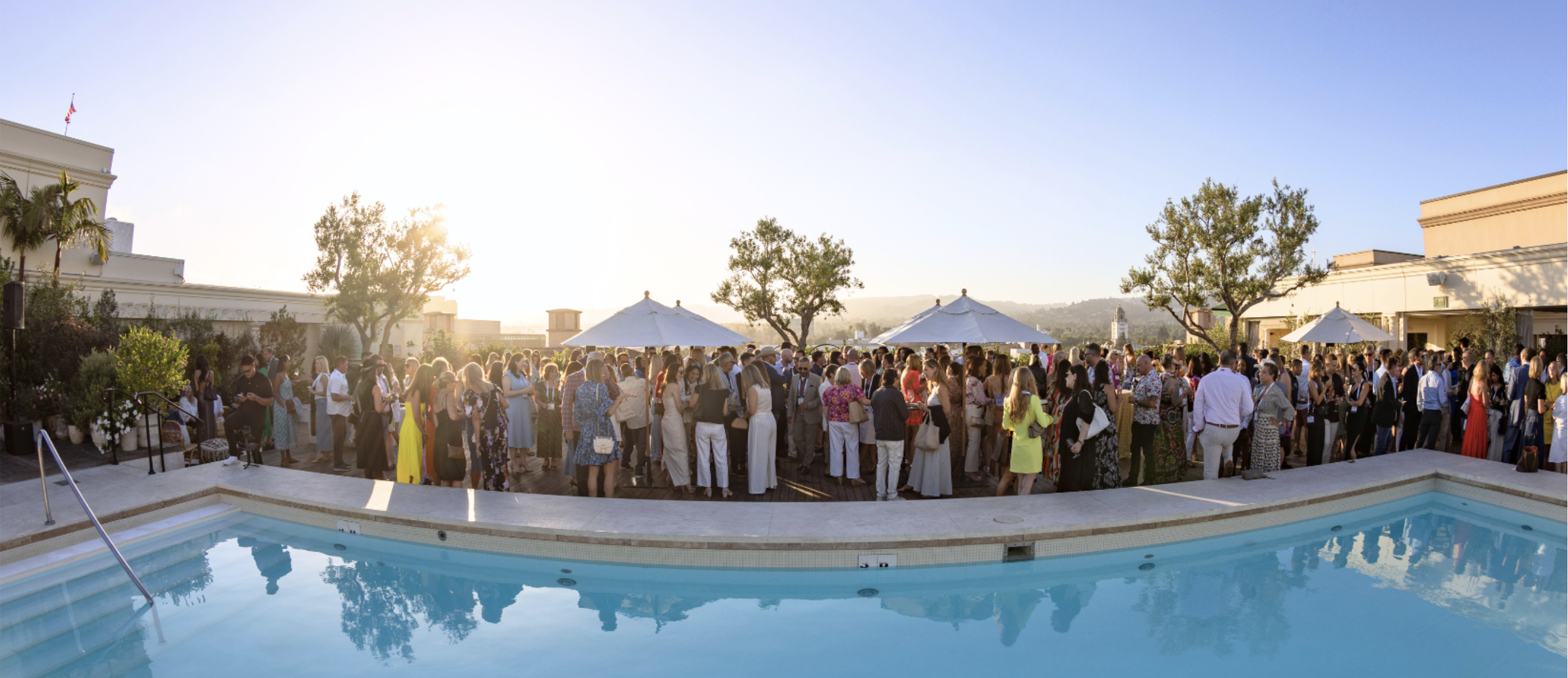 A large group of people gathered outdoors at a social event on a rooftop terrace with a swimming pool in the foreground, sun setting in the background, with trees and buildings visible.
