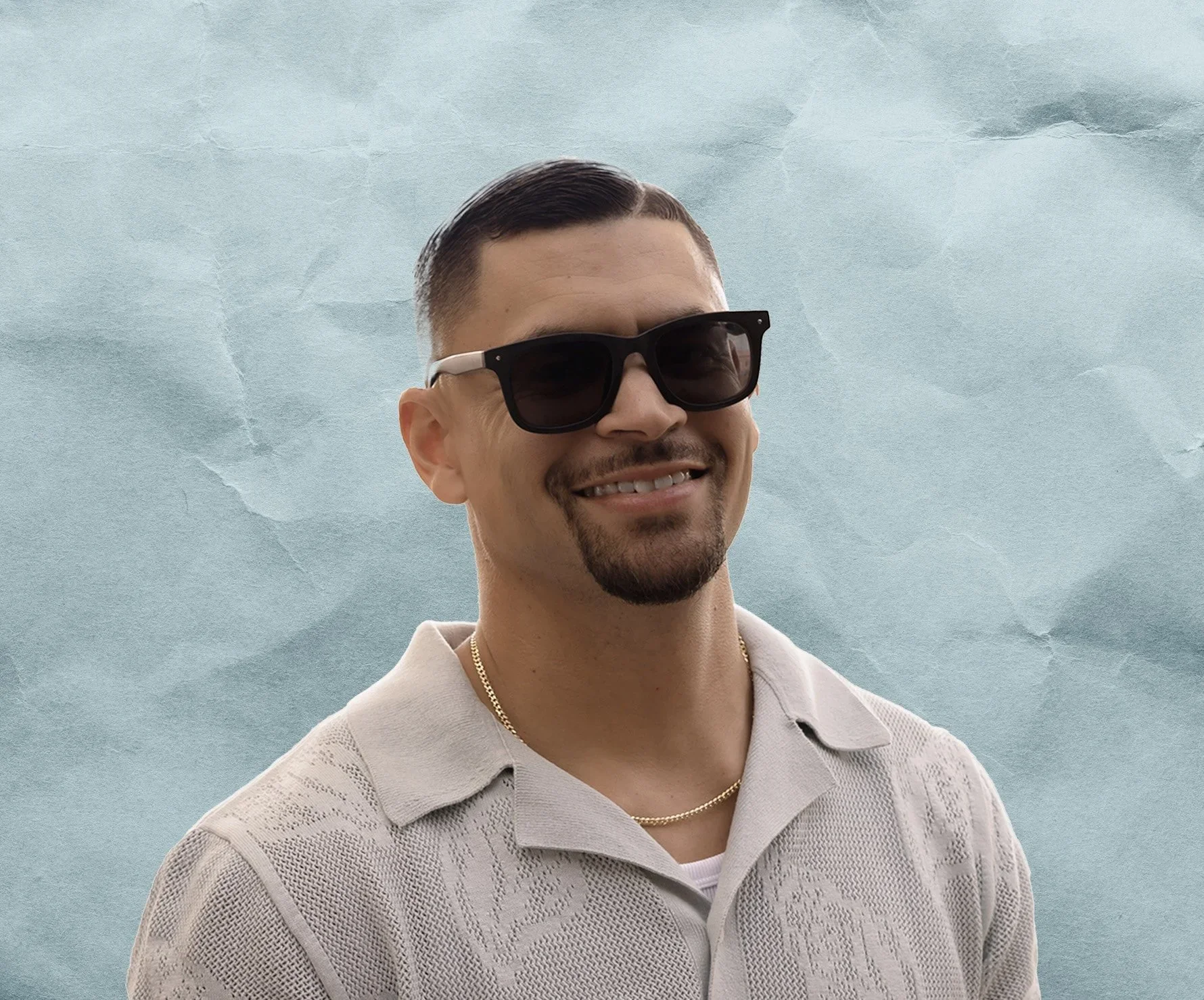 A man smiling, wearing black sunglasses, a beige knitted polo shirt, and a gold chain necklace, standing against a textured light blue background.