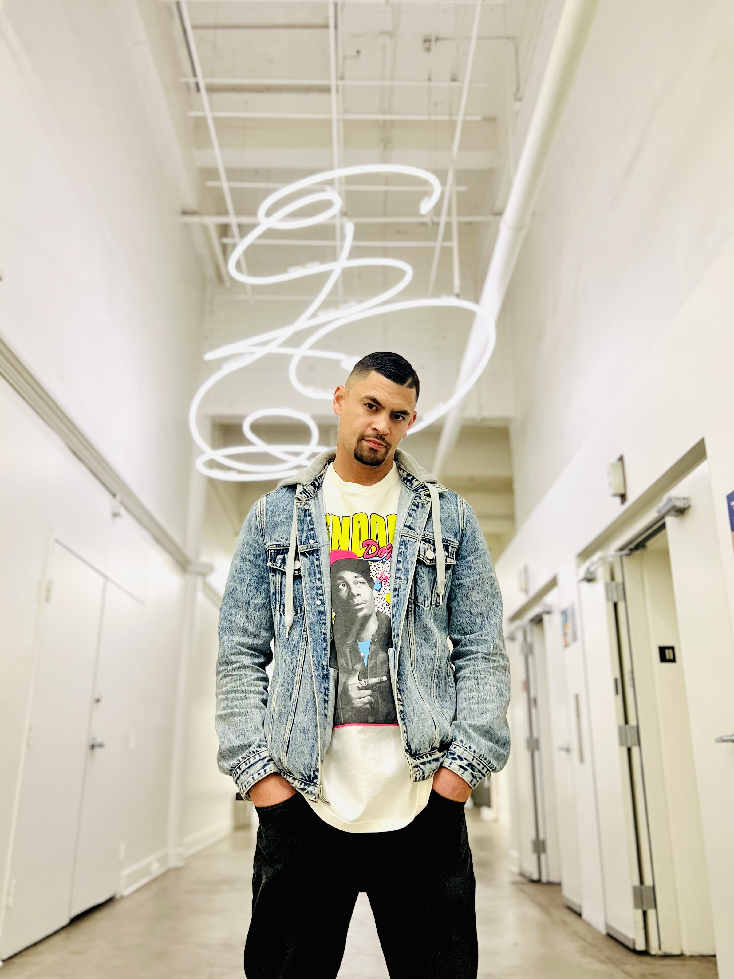 Young man standing in a hallway with white walls and a neon light ceiling art piece.