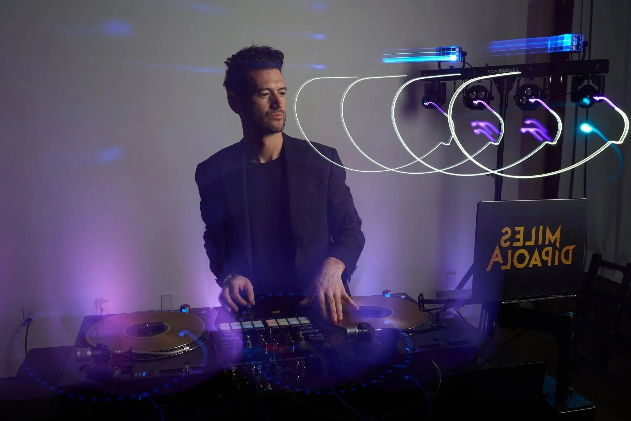 A DJ with dark hair and beard, wearing a black jacket, is performing with turntables and DJ equipment, using a focus light, in a dimly lit room with neon light effects.