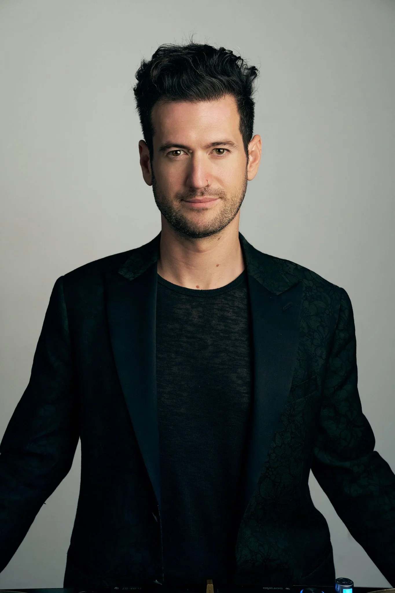 A man with dark, styled hair and a short beard, wearing a black blazer and t-shirt, standing against a plain gray background.