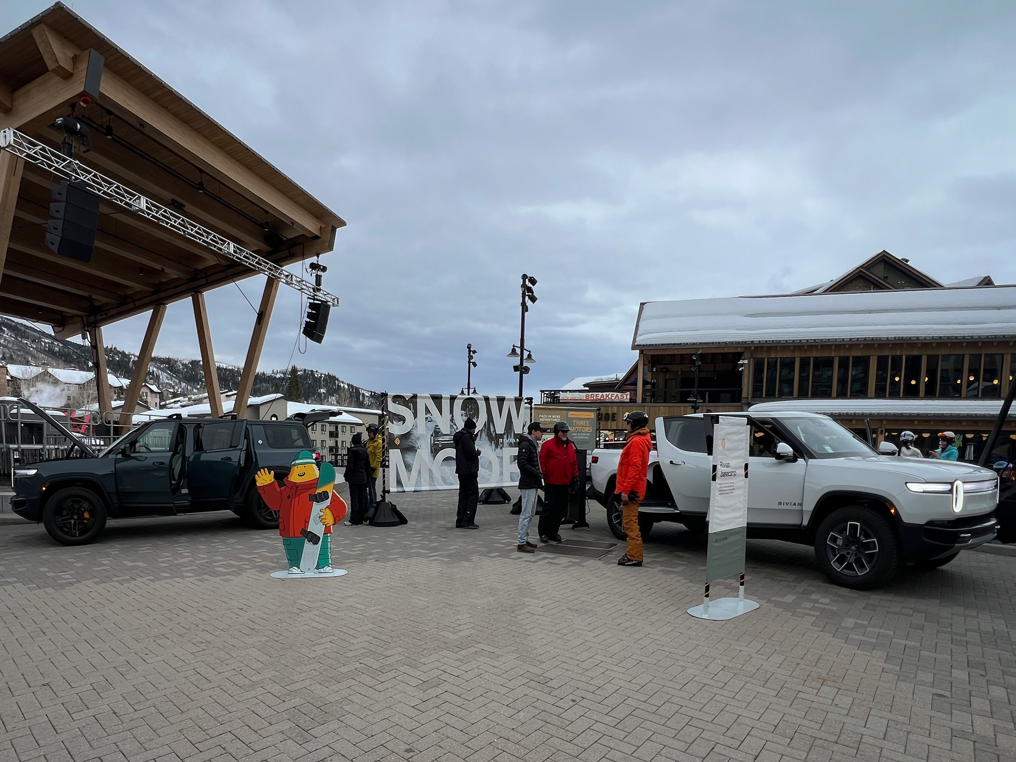 An outdoor market area with several parked cars and people standing around. There is a large sign that says "SNOW" and a cardboard cutout of a person wearing winter clothing. The background features buildings with snow-covered roofs and cloudy skies.