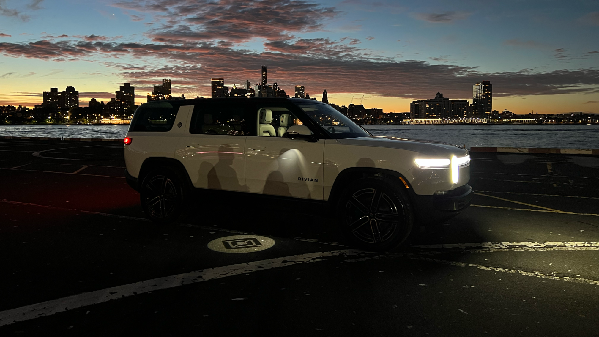 A white Rivian electric SUV with headlights on, parked in a lot at sunset with a city skyline and river in the background.