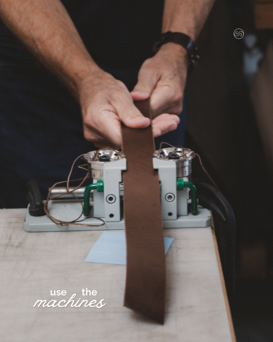 Person operating a leather strap cutting machine on a wooden table.