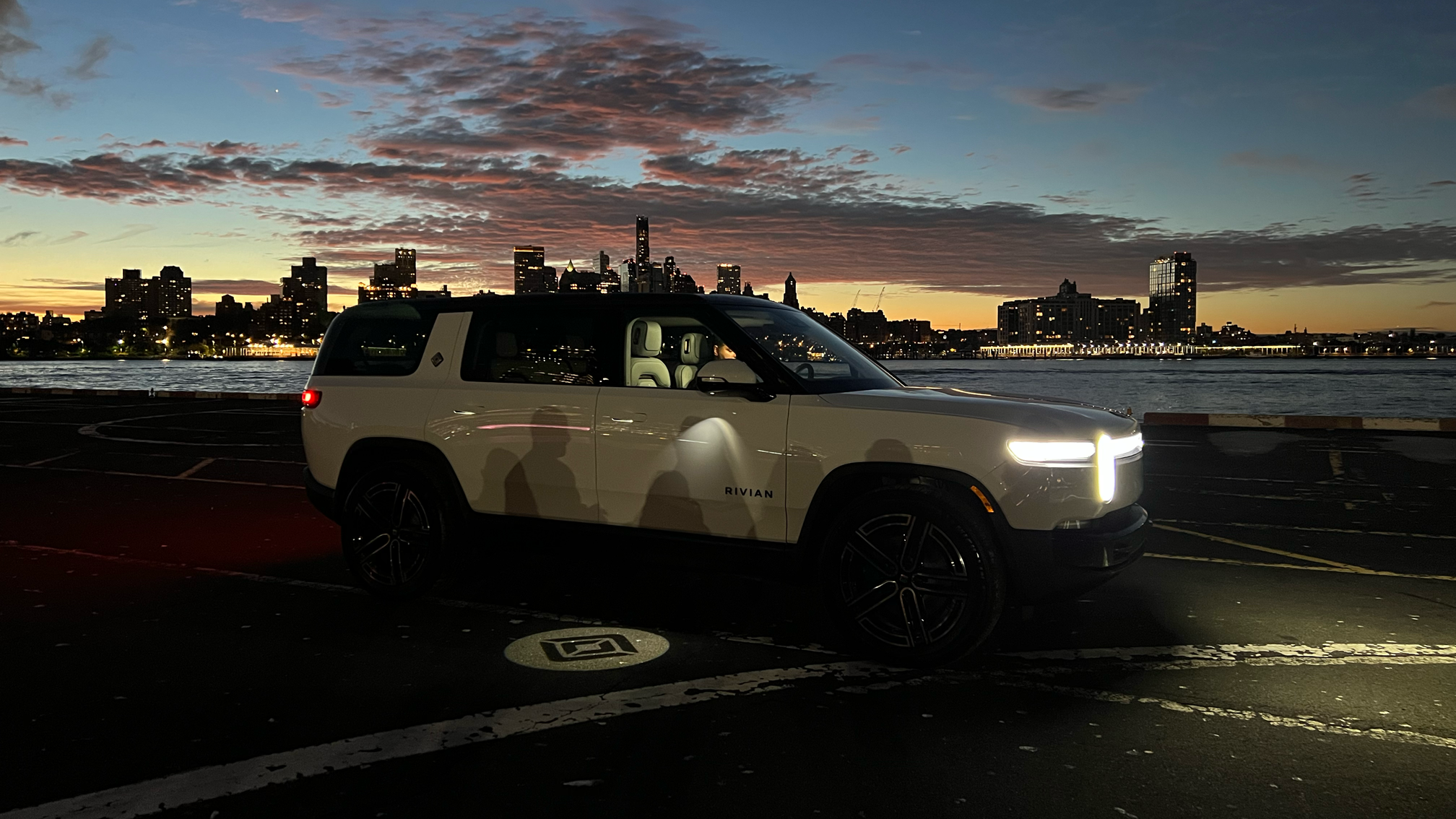 A white Rivian electric SUV parked in a lot near a body of water at sunset, with a city skyline and partly cloudy sky in the background.