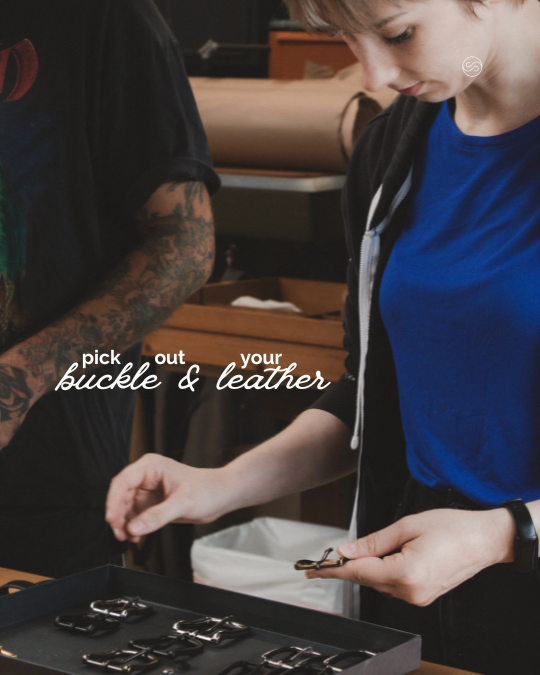 A person with a tattooed arm holding a buckle, inspecting a leather strap on a table with metal hardware, while another person observes.