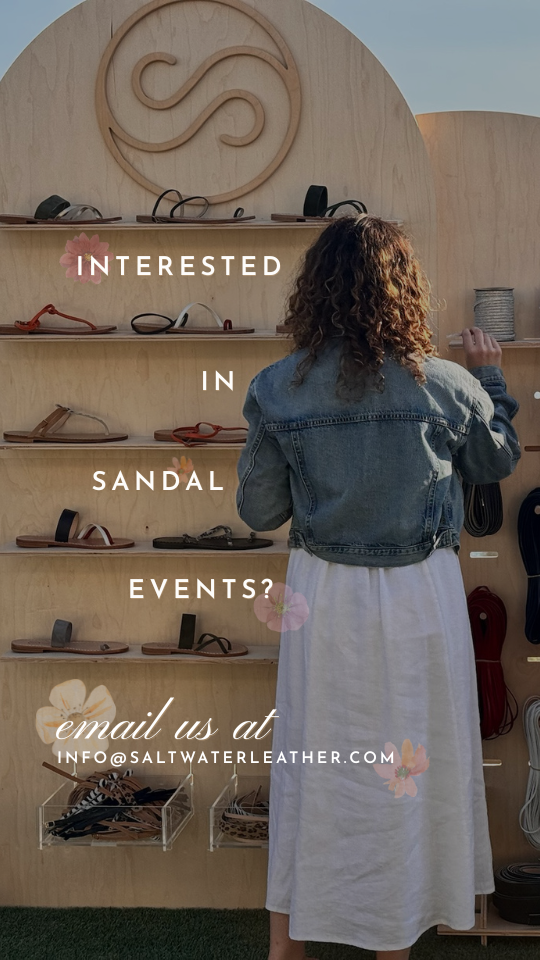 A woman with curly hair wearing a denim jacket and white skirt shopping for sandals at an outdoor booth with shelves of sandals and a Salt Water Leather sign overhead.