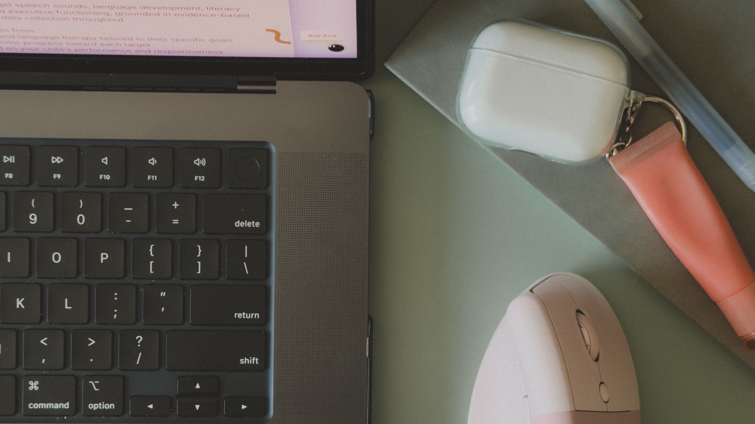Top view of a workspace with a laptop keyboard, a smartphone displaying text, a white AirPods case, a pink lipstick, a pen, a closed notebook, and a pink computer mouse on a light green surface.