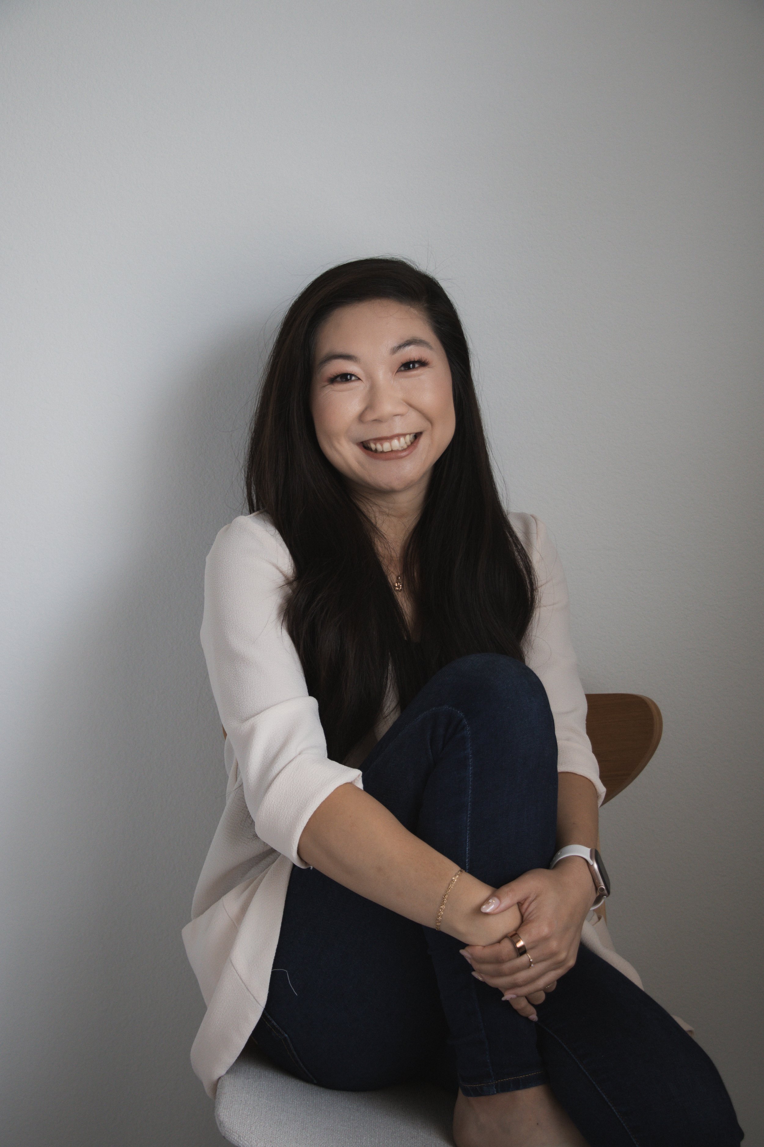 A woman with long dark hair smiling, sitting on a chair against a plain gray wall, wearing a white top and dark jeans, with her arms crossed over her knee.