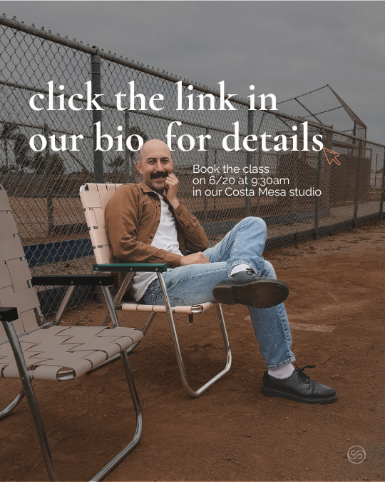A smiling man with a beard, wearing a brown jacket, white shirt, and jeans, sitting on a folding chair on a dirt baseball field next to a chain-link fence; text overlay promoting class booking at Costa Mesa studio.
