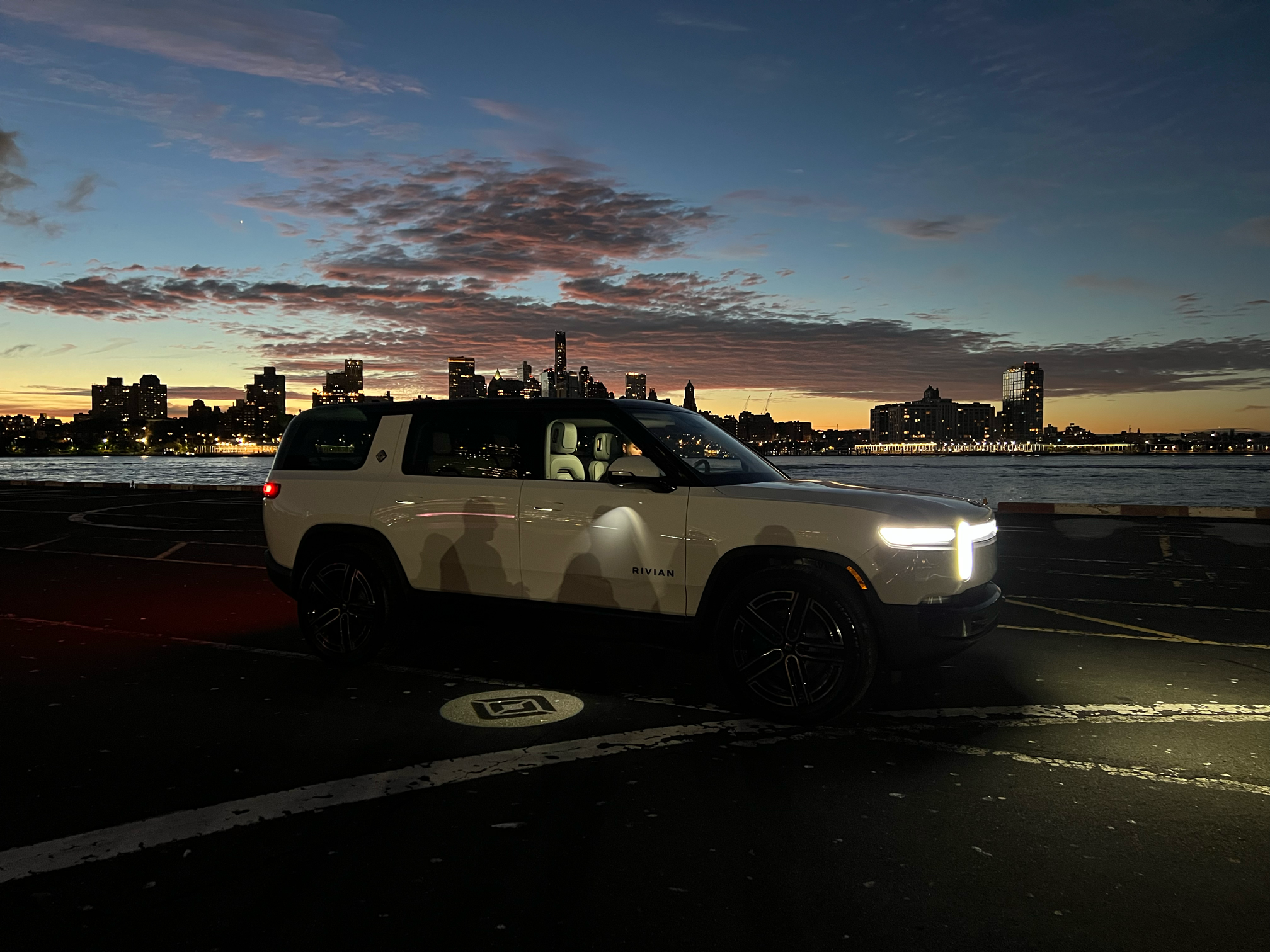 A white SUV with the label 'RIVIAN' on the side is parked on a dark parking lot during sunset, with a city skyline and a partly cloudy sky in the background.