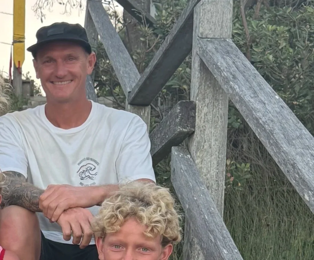 A smiling man with light skin, wearing a black cap and white t-shirt, sitting outdoors on wooden stairs next to a young boy with curly blonde hair and light skin, in a natural setting with greenery.