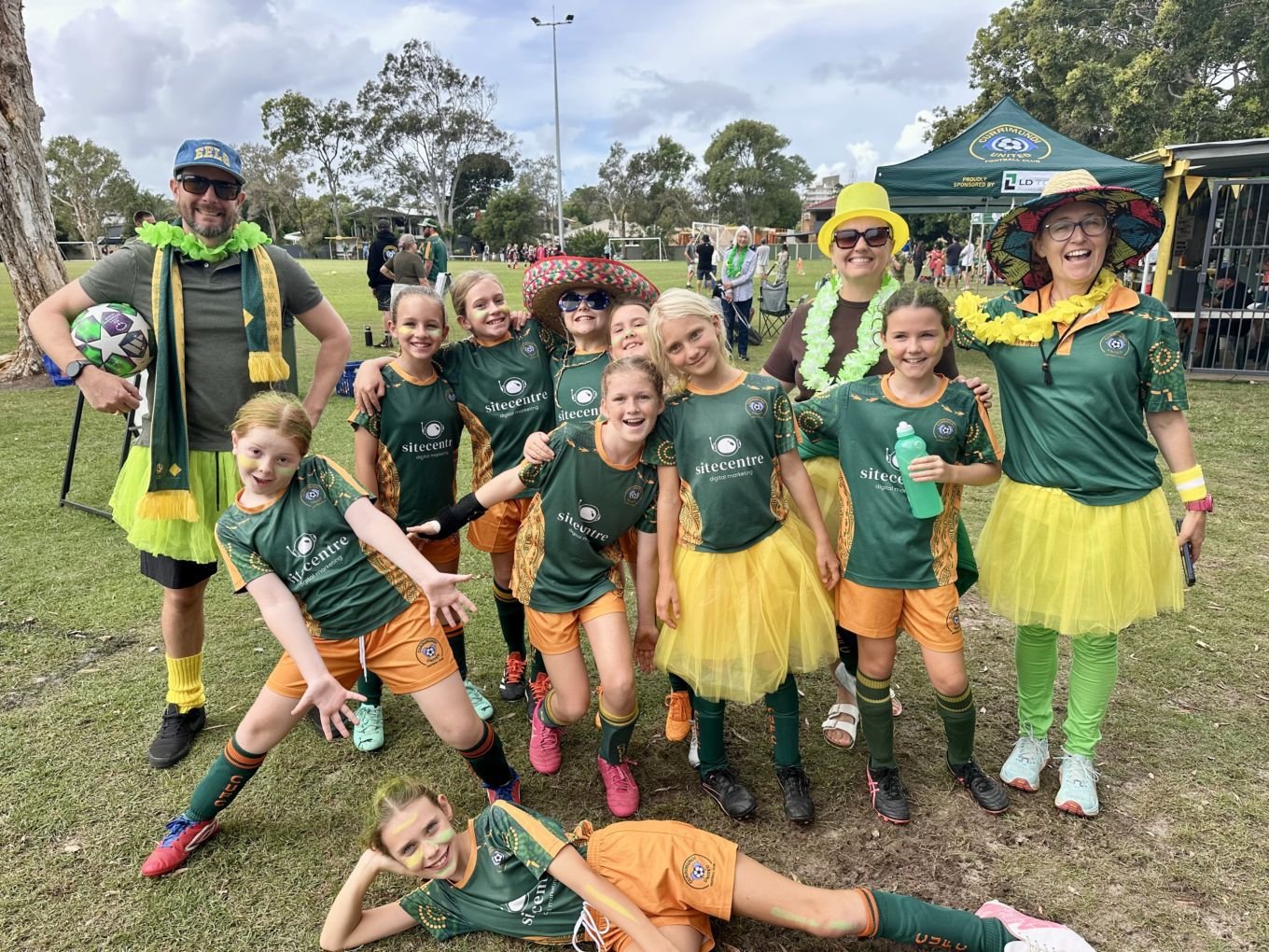 A group of children and adults dressed in sports uniforms with green and yellow colors, wearing hats and accessories, celebrating on a grassy field during a sporting event or festival.