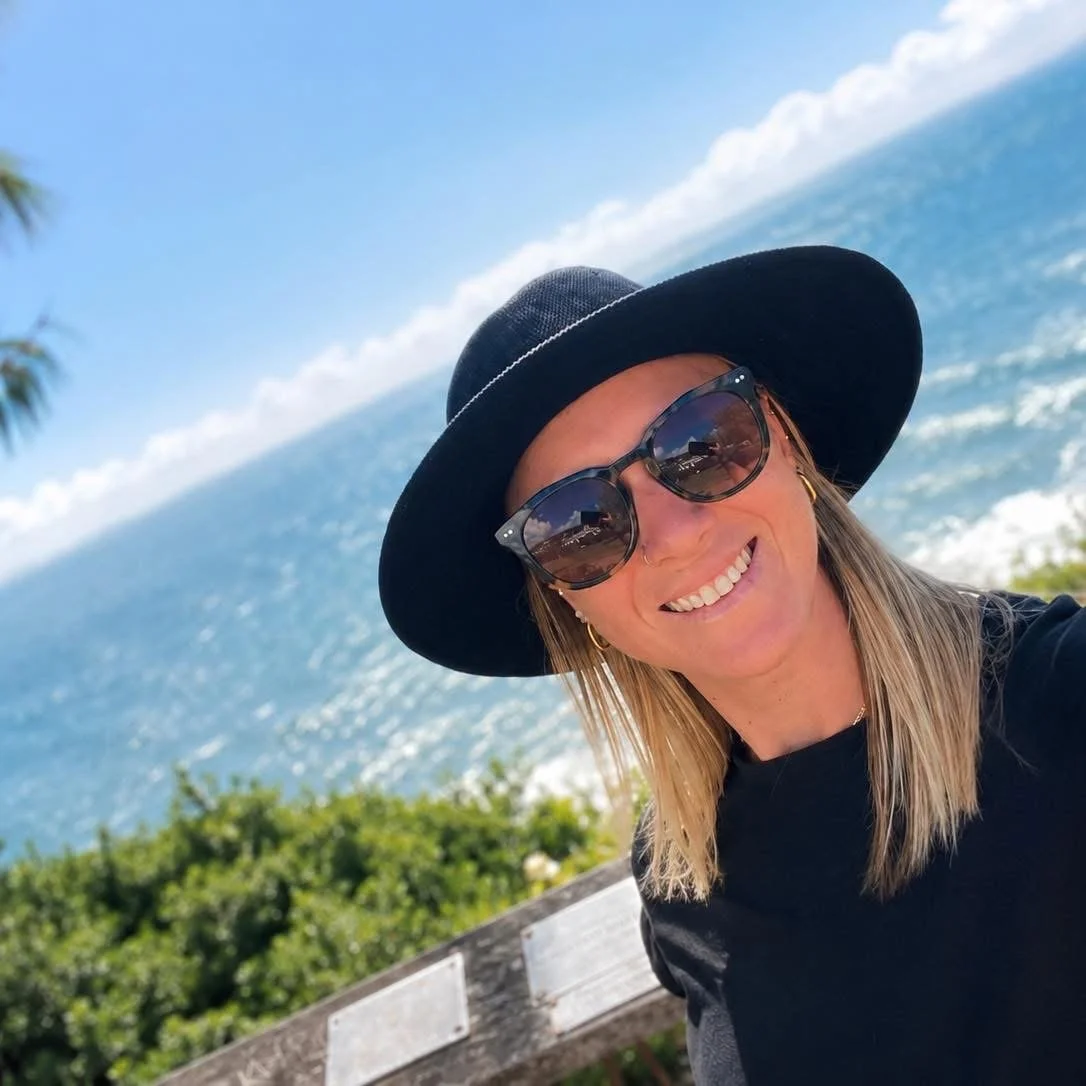 A woman smiling wearing sunglasses and a black wide-brimmed hat, with a view of the ocean and blue sky in the background.
