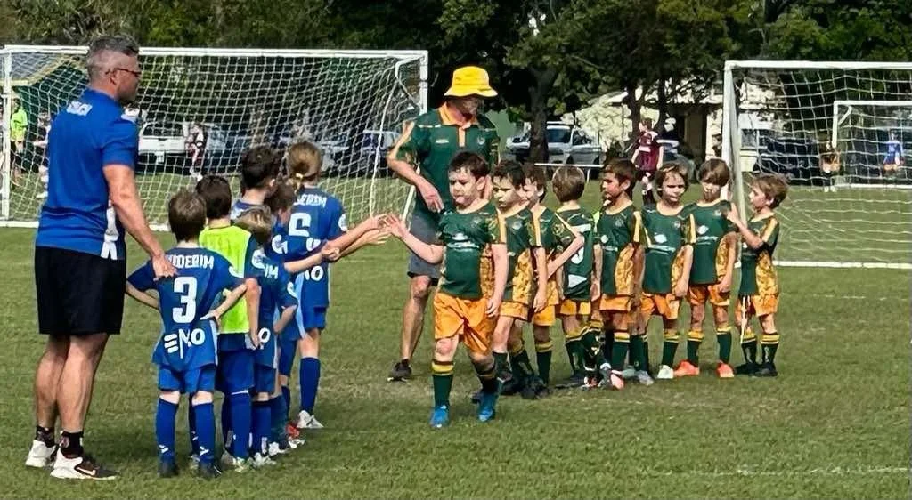 Youth soccer teams in jerseys shaking hands on a field after a game, with coaches present, and soccer goals and trees in the background.