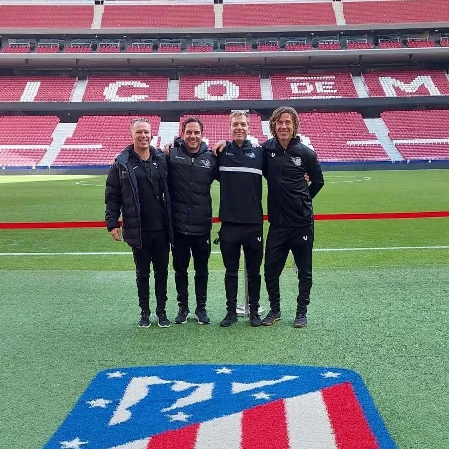 Four men standing on a soccer field in a stadium, smiling with their arms around each other. They are dressed in sports tracksuits. In the background, stadium seats spell out "IGOR DE M" with red and white colors. The ground features a large logo of a sports team or organization.