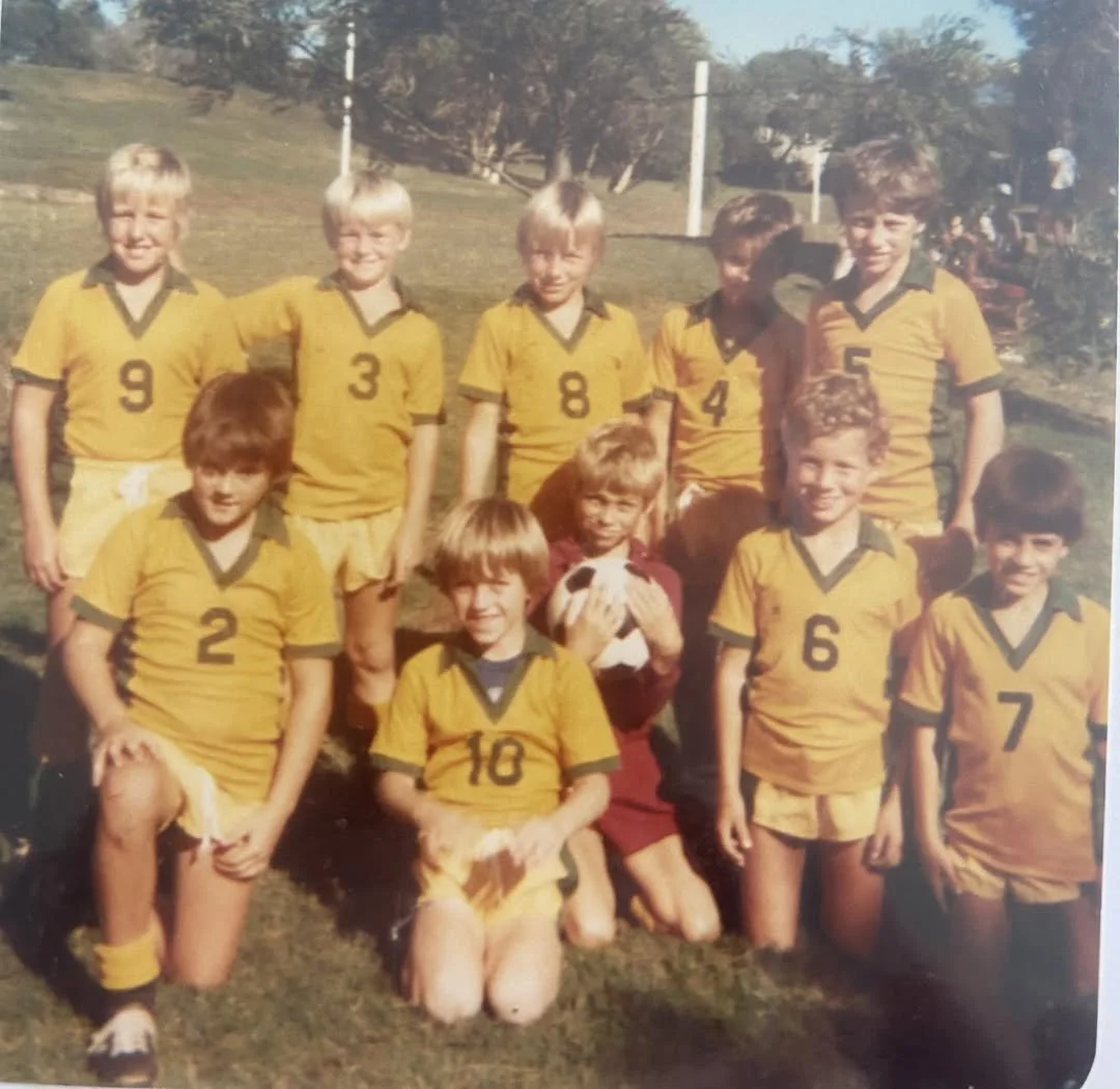 Group of young boys in yellow and green sports uniforms posing outdoors, some kneeling and some standing, with a girl holding a white and brown dog in the center.