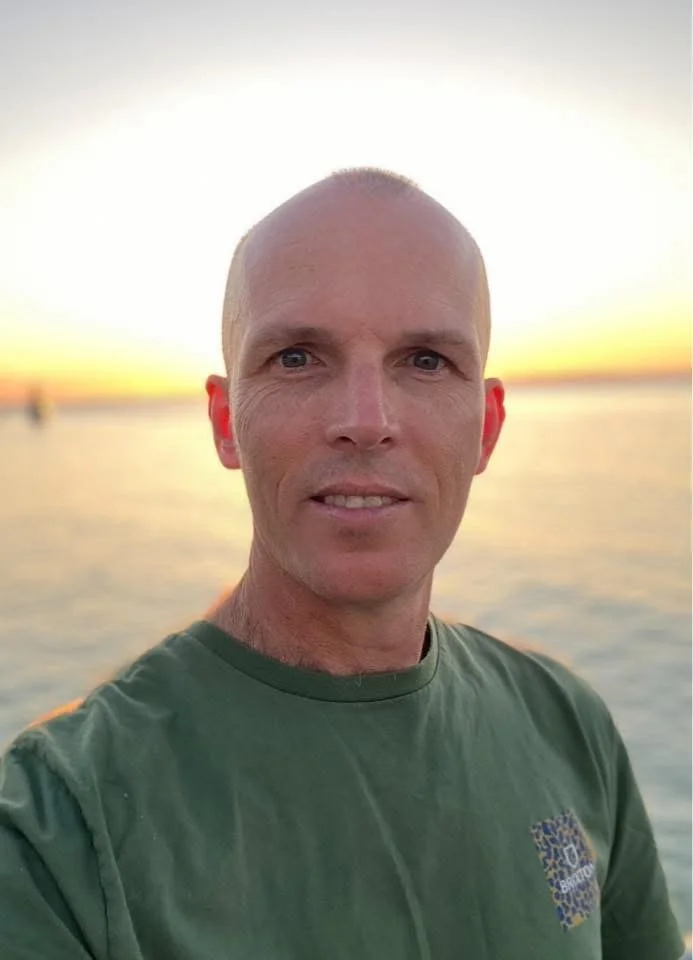 A man with a shaved head taking a selfie at sunset by the water.