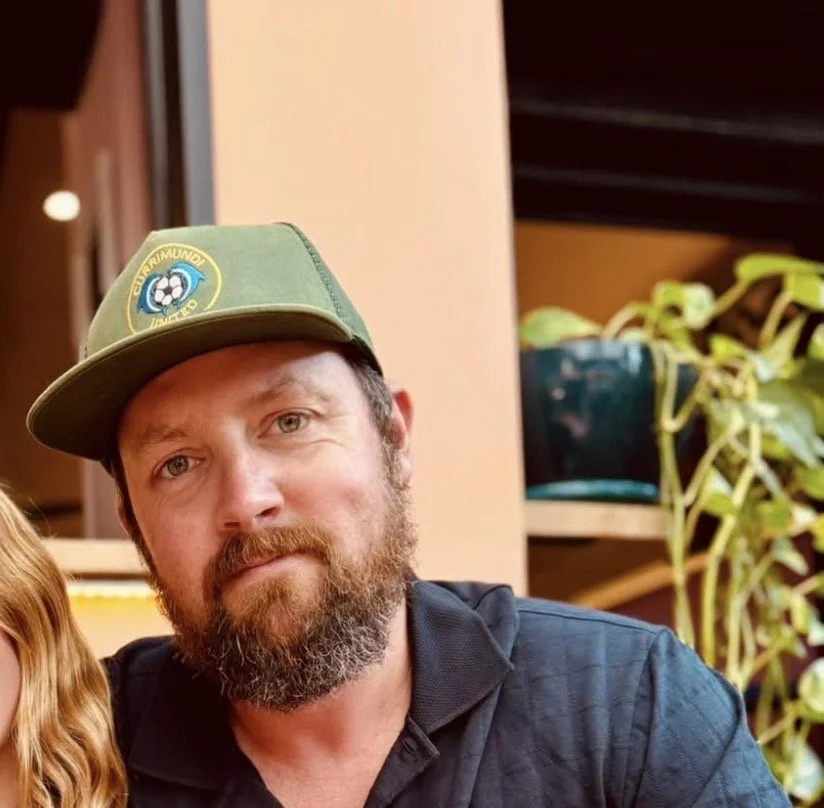 A man with a beard, wearing a green cap with a soccer logo and a black shirt, sitting indoors with a potted plant and furniture in the background.