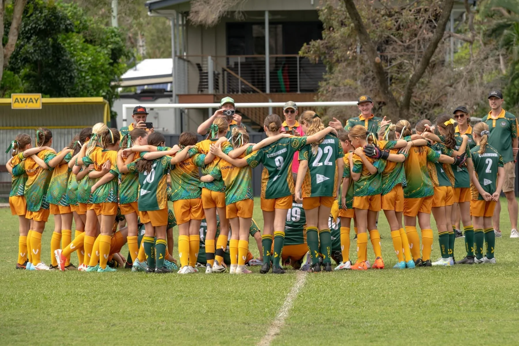 A group of young female soccer players in green and yellow uniforms huddle together on a grass field during a match or practice, with some coaches and spectators watching.